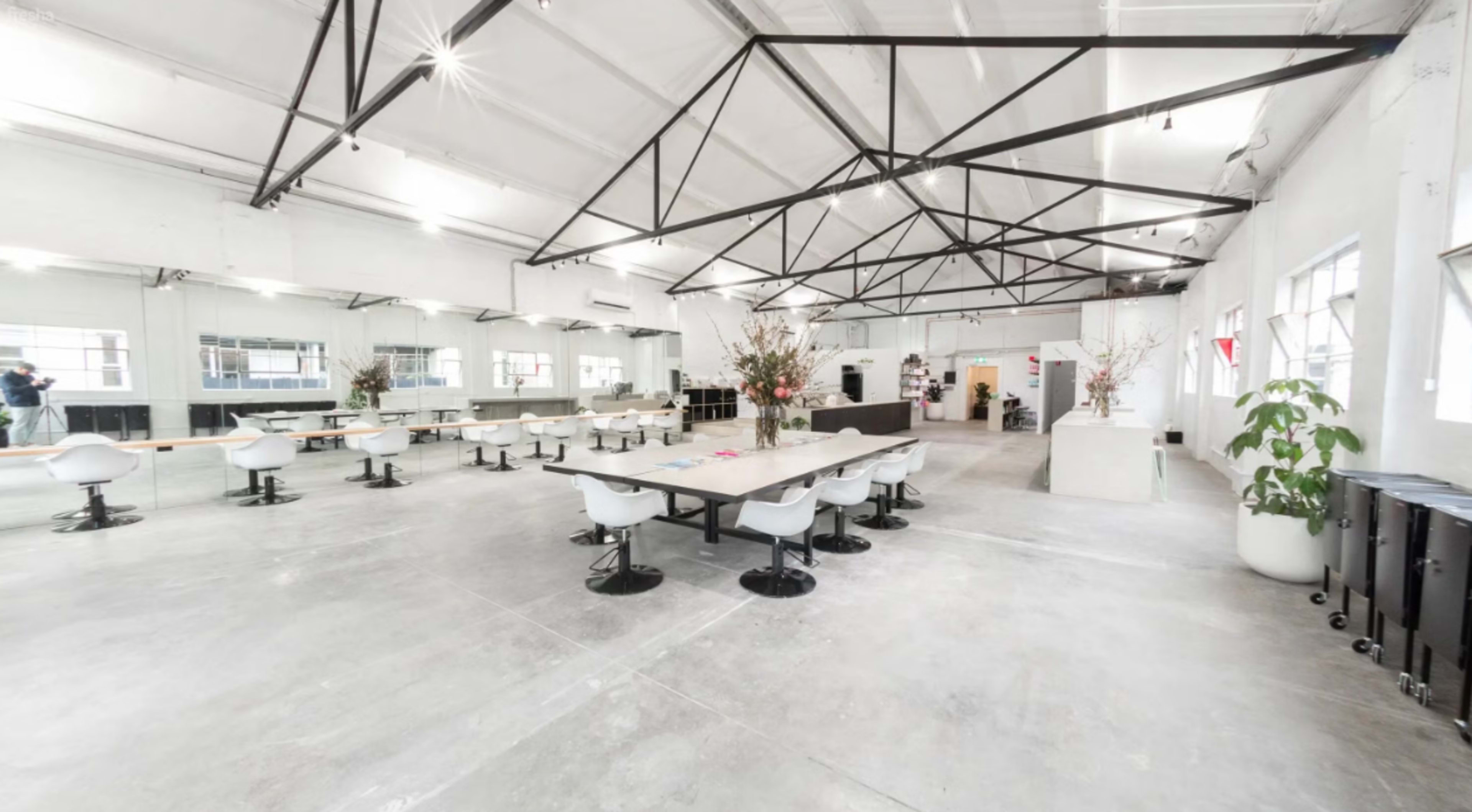 The image shows a spacious, modern event space with a long table and white chairs, surrounded by large windows and minimalist decor.