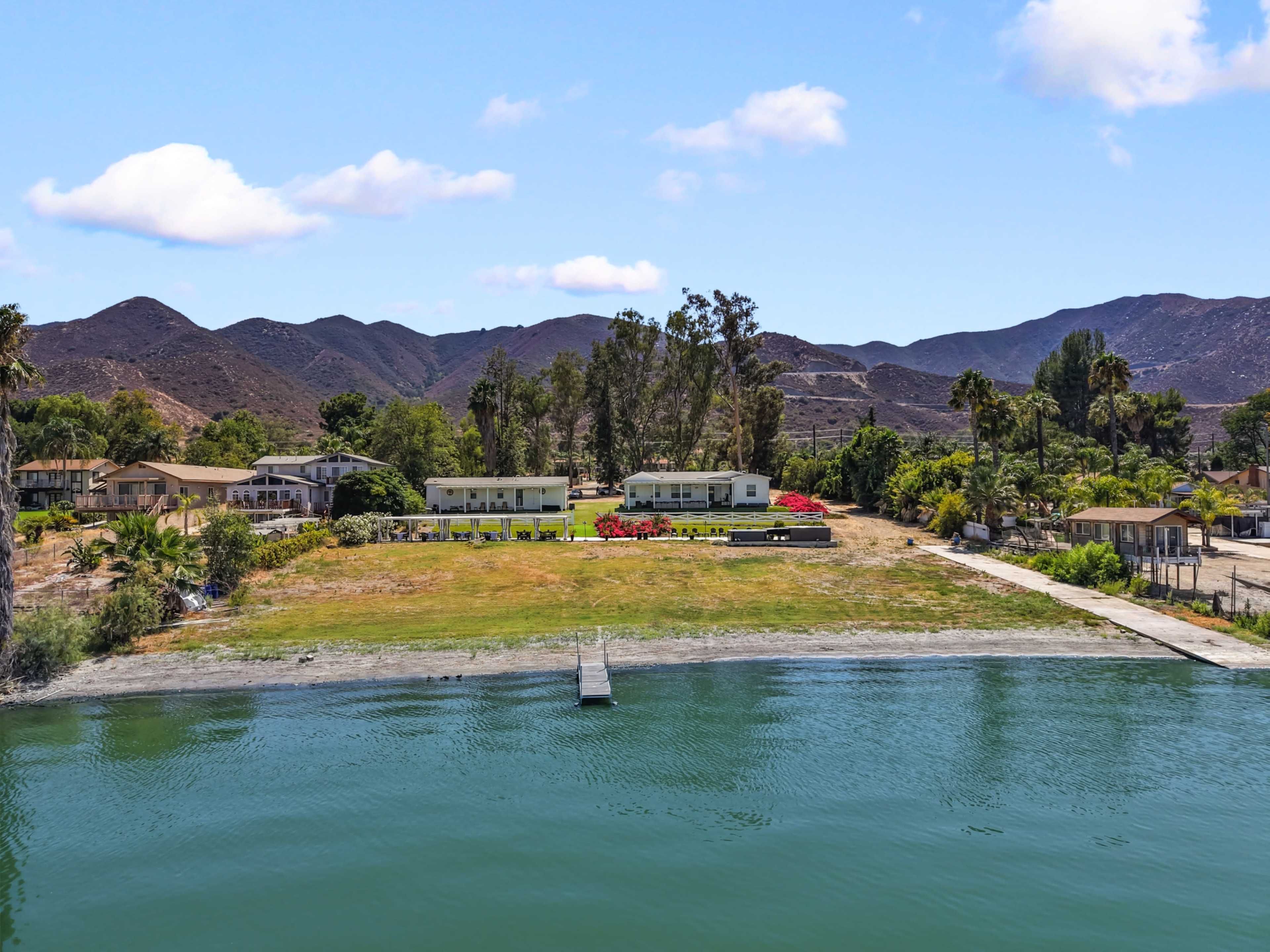 The image shows a lakeside property with several buildings, a dock extending into the water, and mountains in the background.