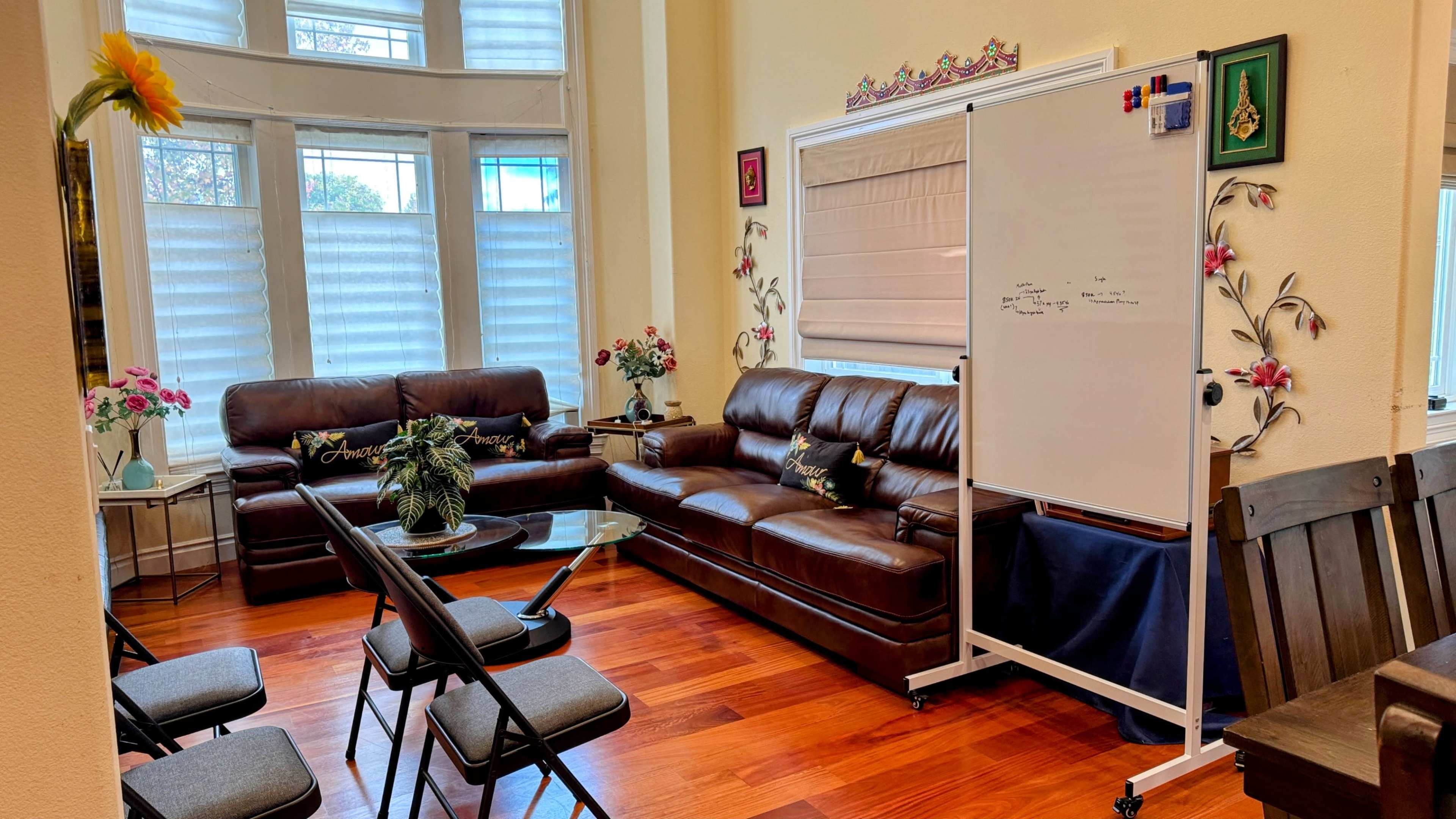 The image shows a cozy meeting room with brown leather couches, a small glass table, and a whiteboard positioned against a wall, surrounded by light-colored walls and decorative plants.