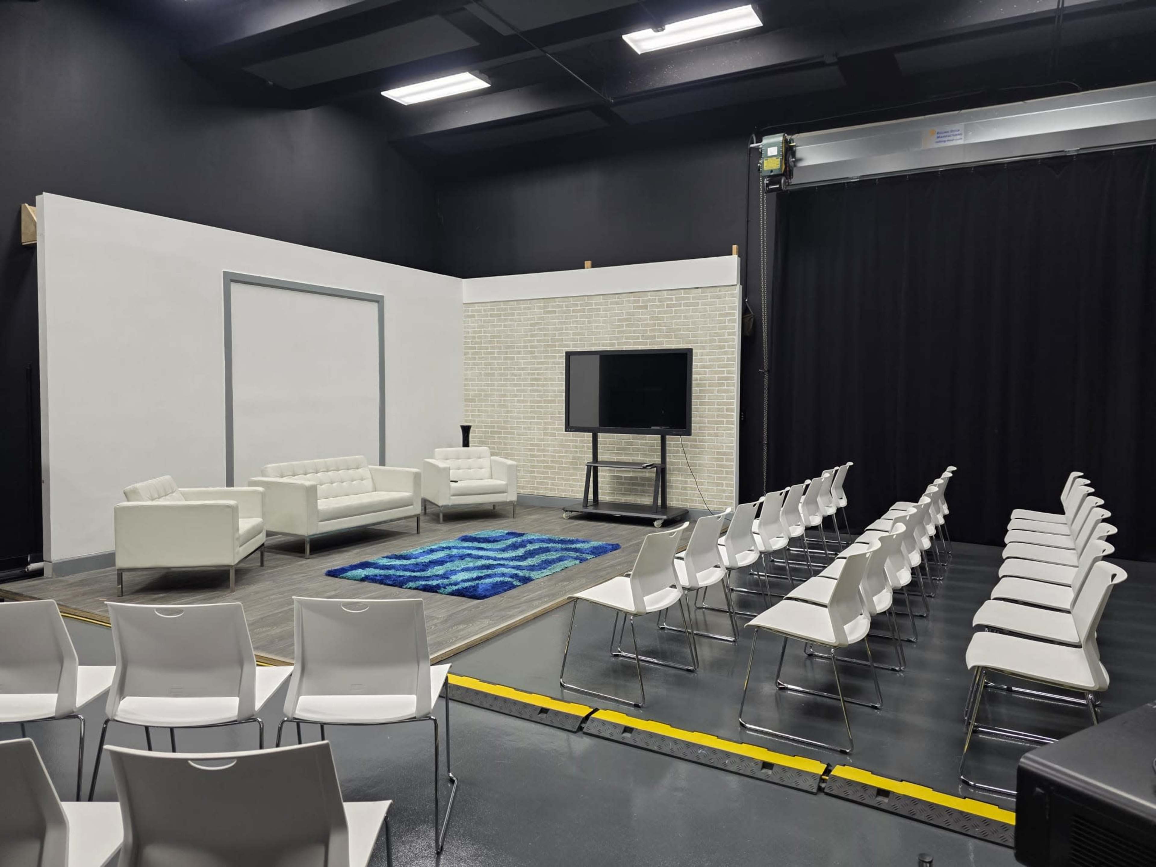 The image shows a sparsely furnished stage area with two white sofas, a television on a stand, and rows of plastic chairs facing the stage.