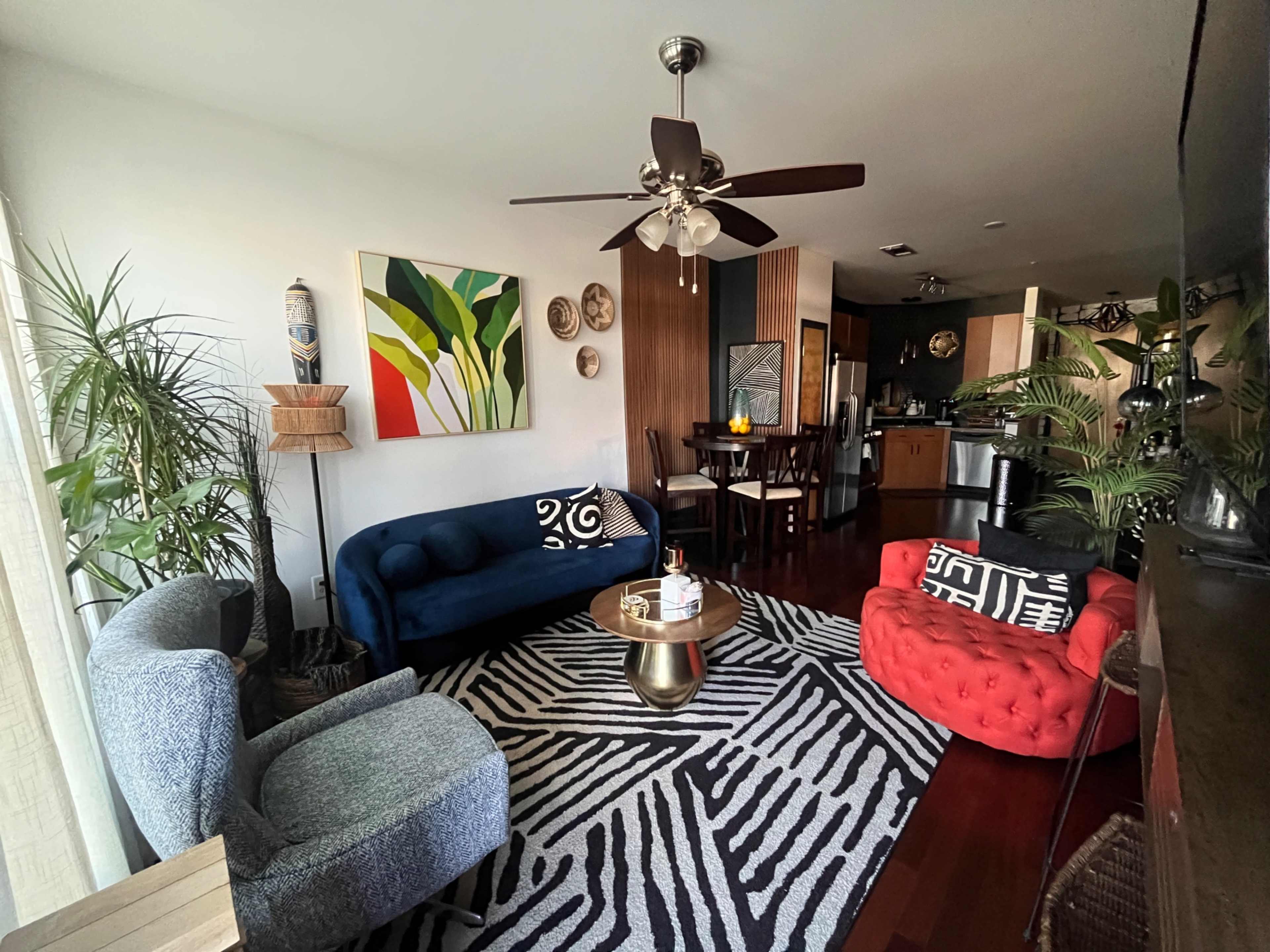 The image shows a cozy living room featuring a blue sofa and a red chair, a round coffee table on a striped rug, and a dining area visible in the background.