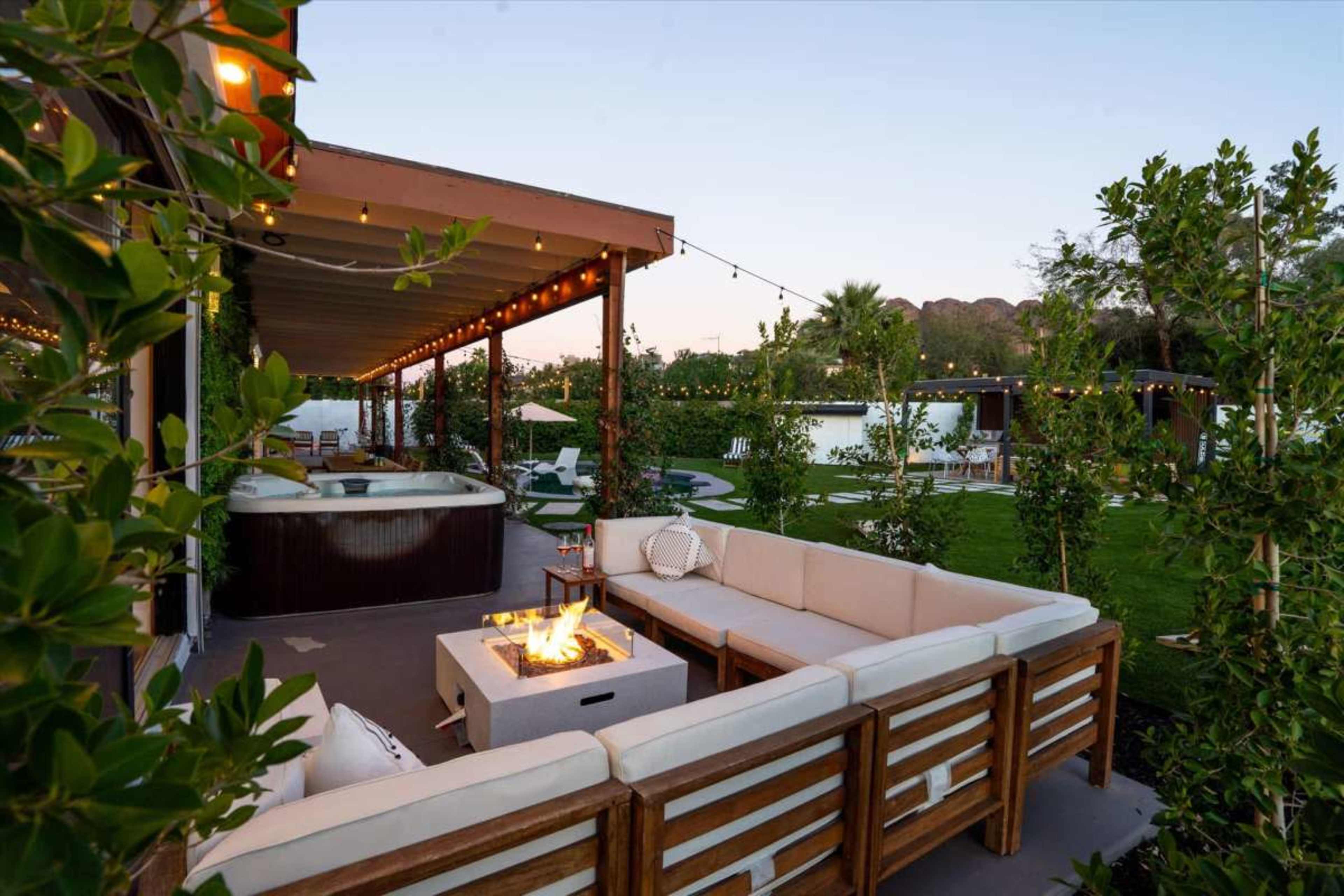 The image shows an outdoor lounge area featuring a fire pit surrounded by a sectional sofa and a hot tub, set in a landscaped garden with string lights overhead.