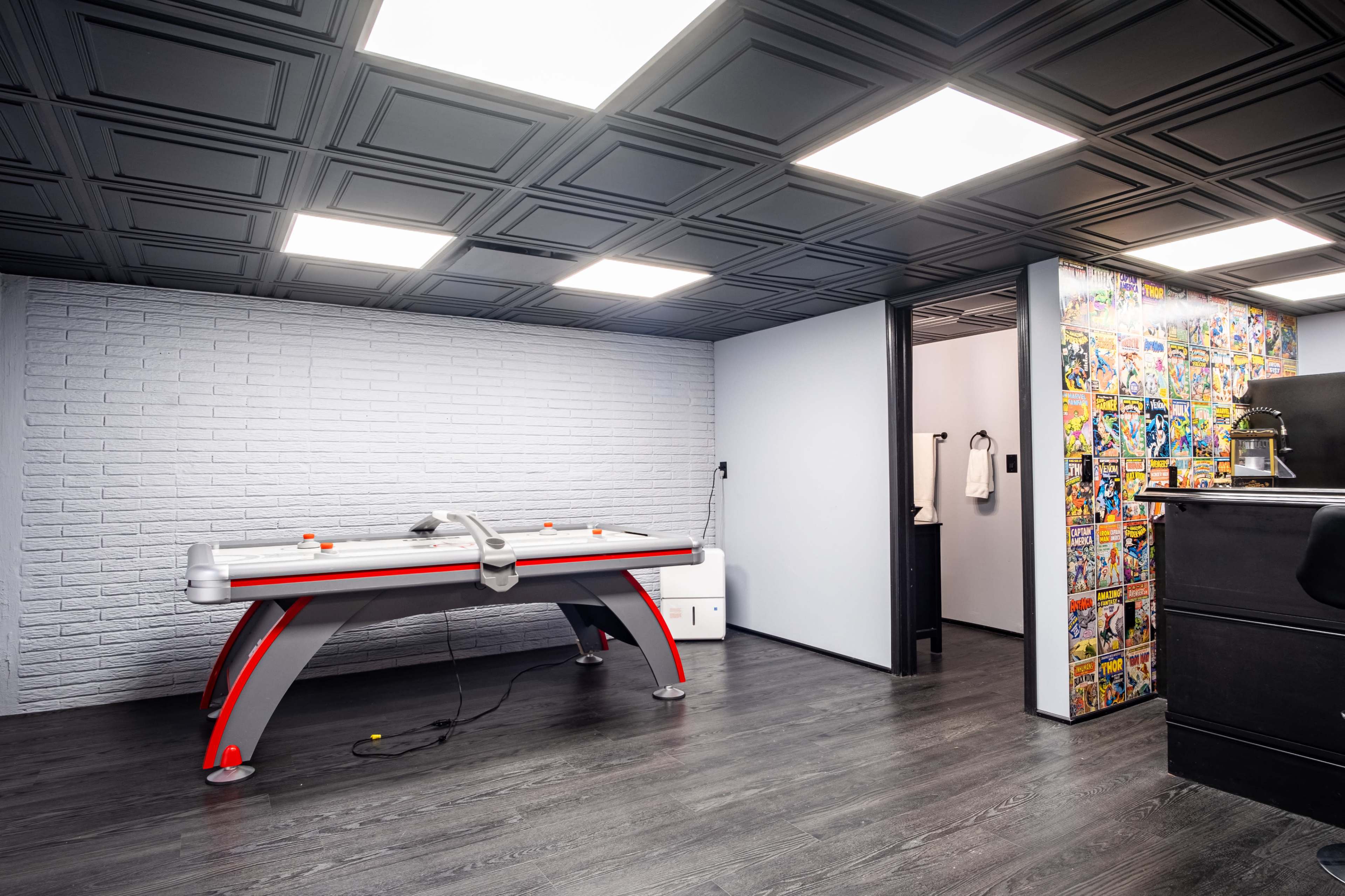 A game room featuring an air hockey table in front of a textured white brick wall, with a door leading to a restroom area and colorful comic book wallpaper on one side.