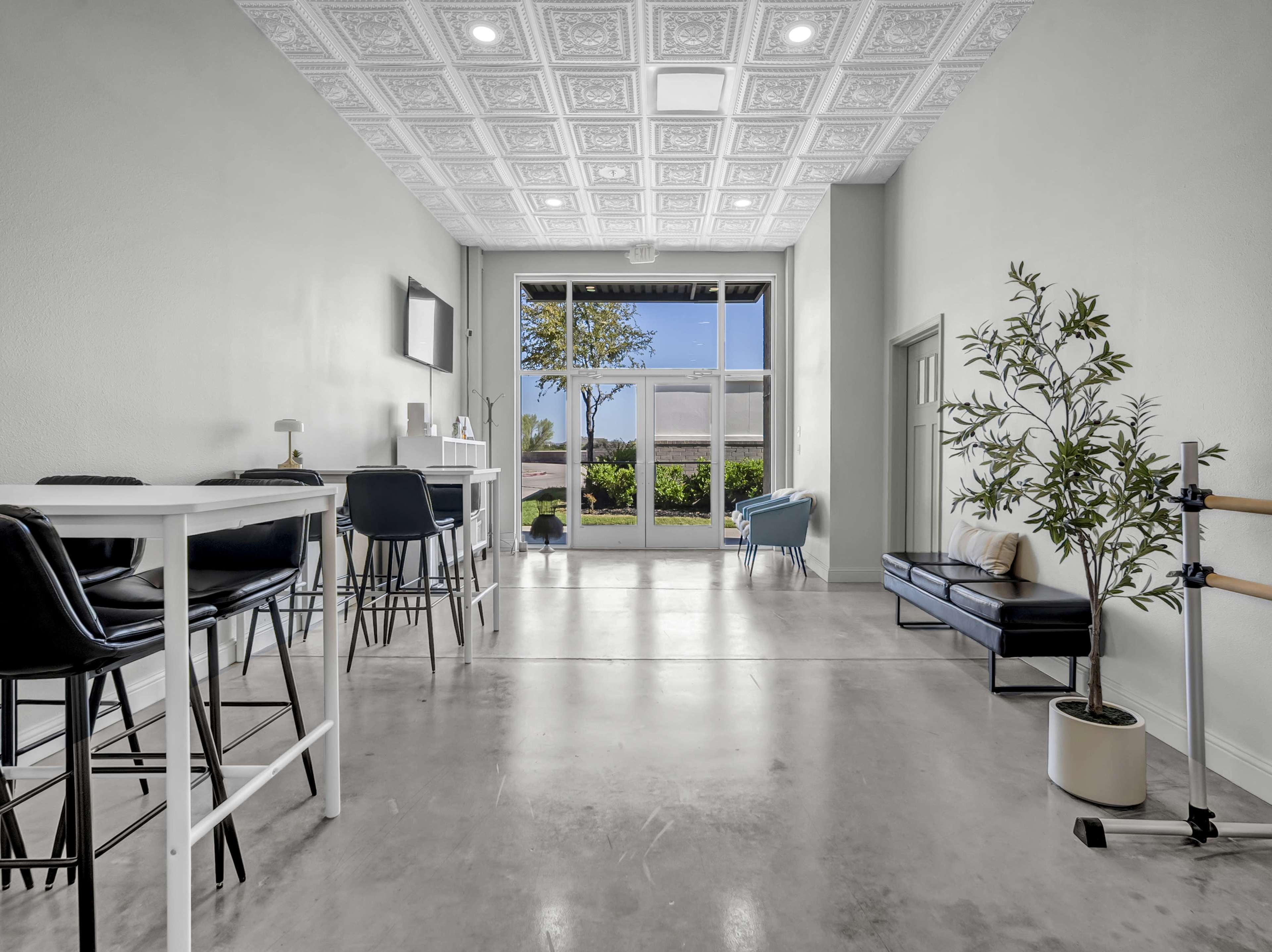The image shows a bright, minimalistic interior space with bar stools, a small table, and large windows that open to a landscaped outdoor area.