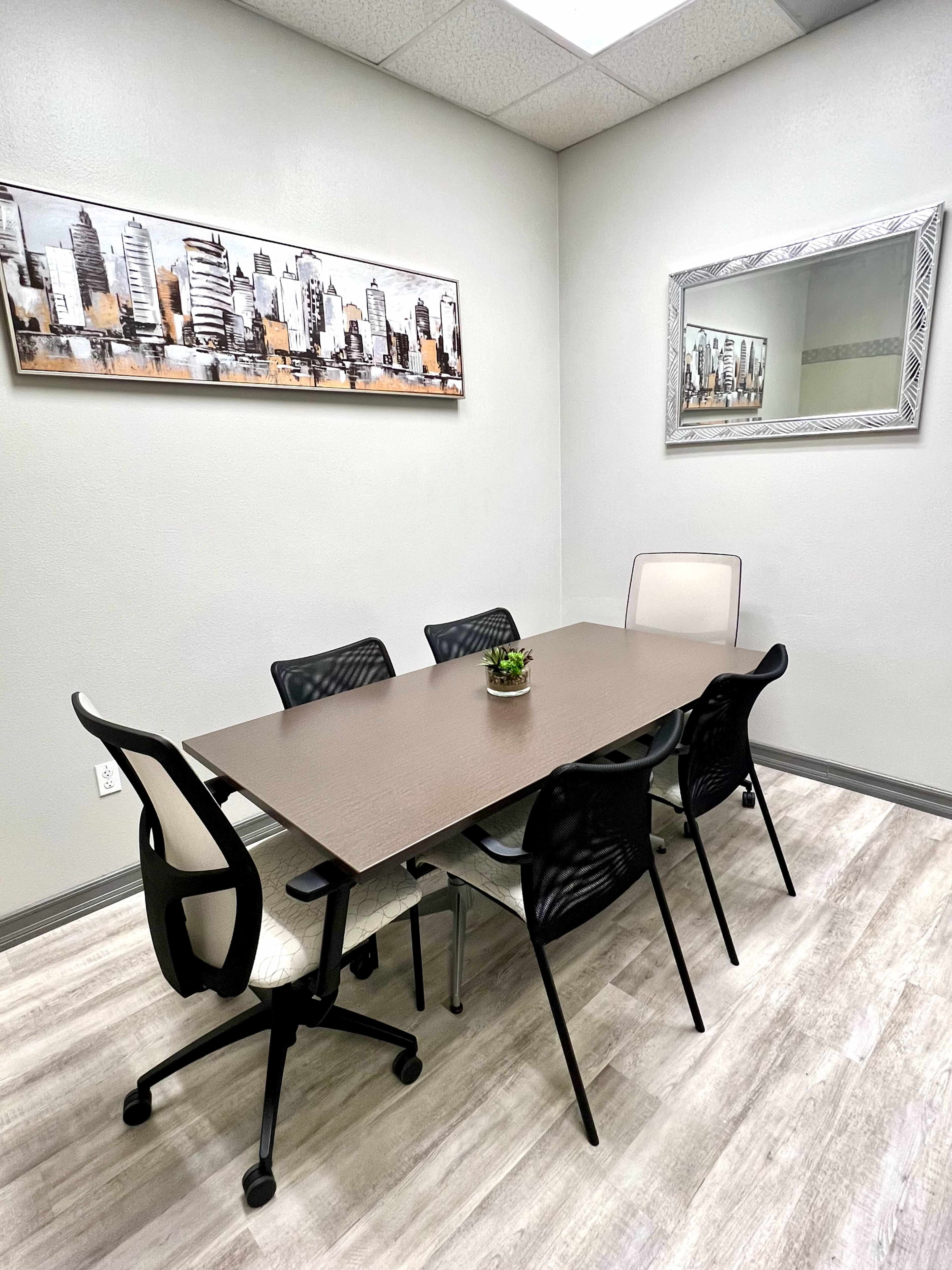 The image shows a small conference room with a rectangular table surrounded by six chairs, a wall-mounted cityscape art piece, and a mirror.