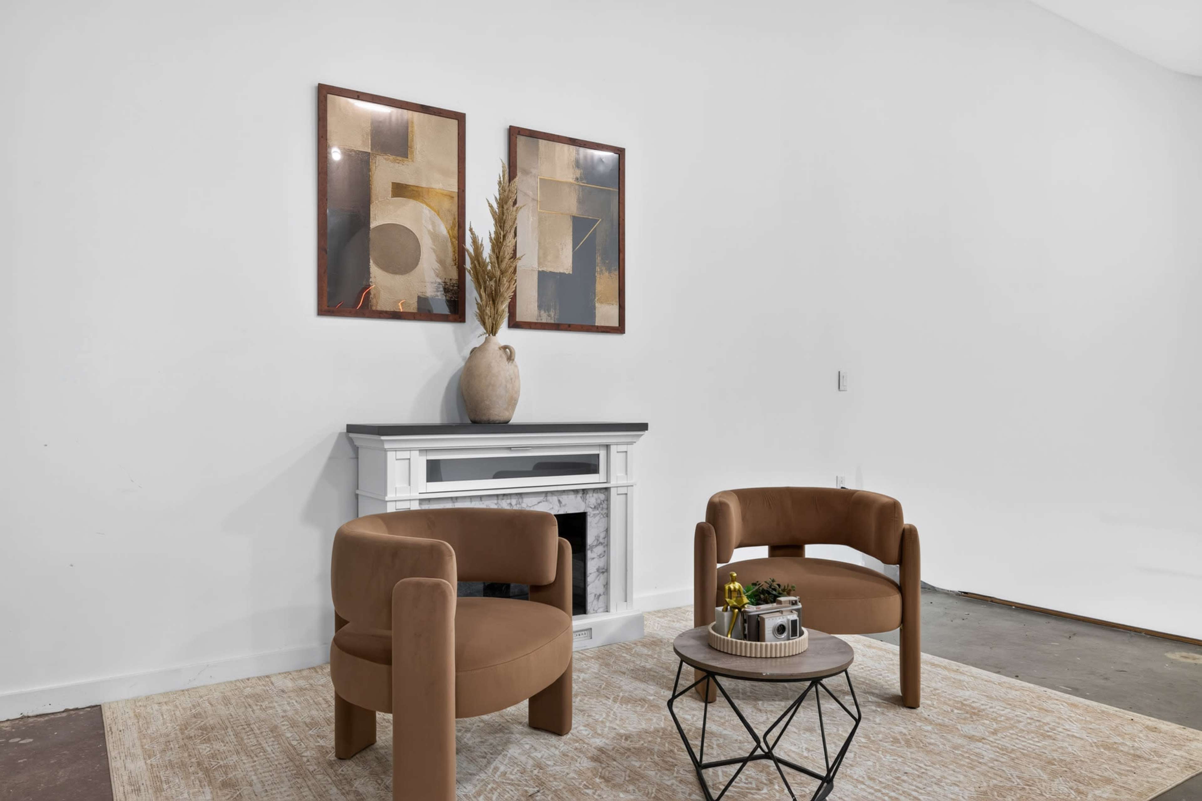 The image features two brown upholstered chairs positioned near a decorative vase on a table in front of a fireplace, with two framed artworks hanging on the wall above.