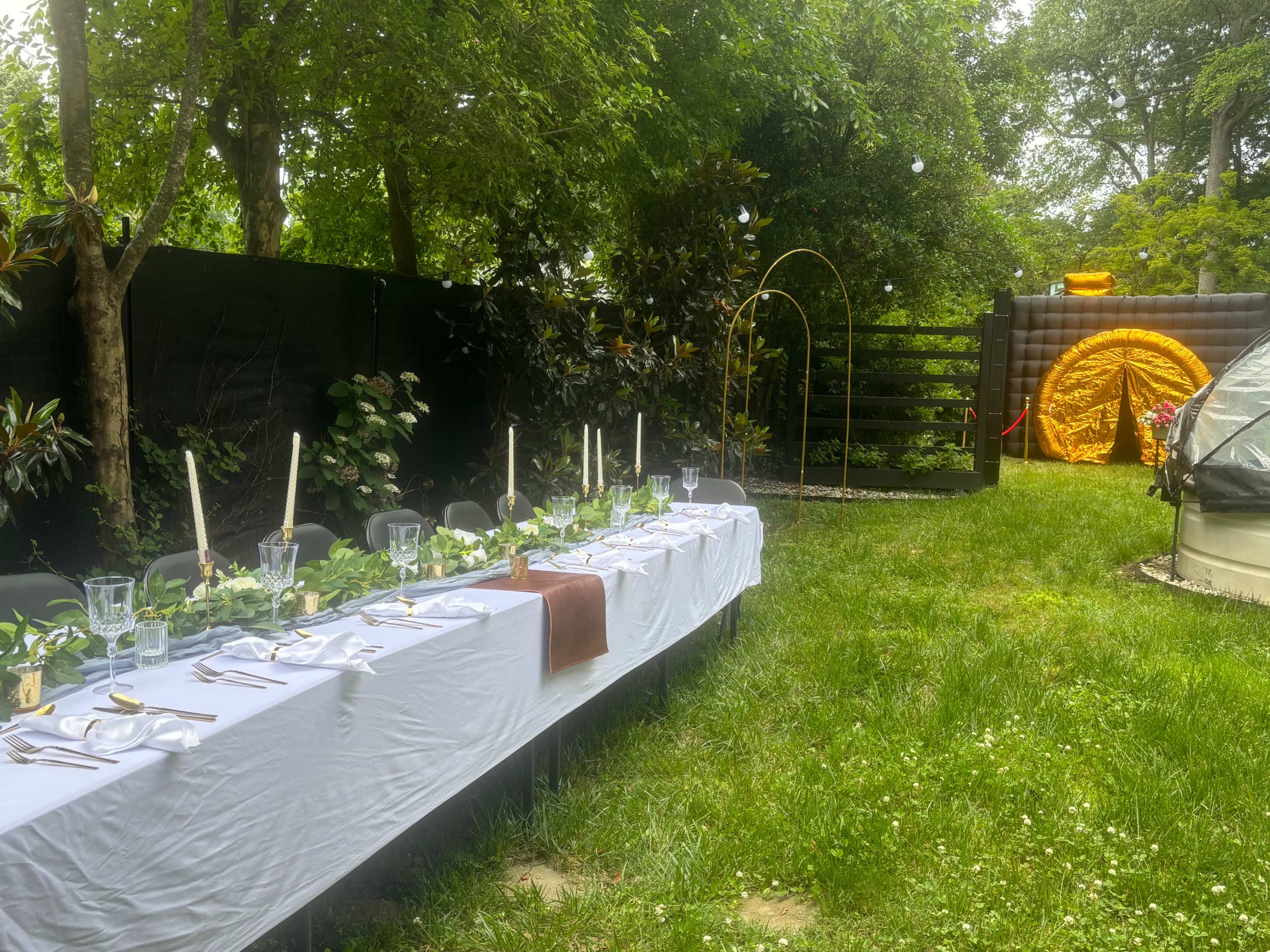 A long banquet table is set up in a garden, adorned with greenery and candles, with a decorative arch and a geodesic dome structure nearby.