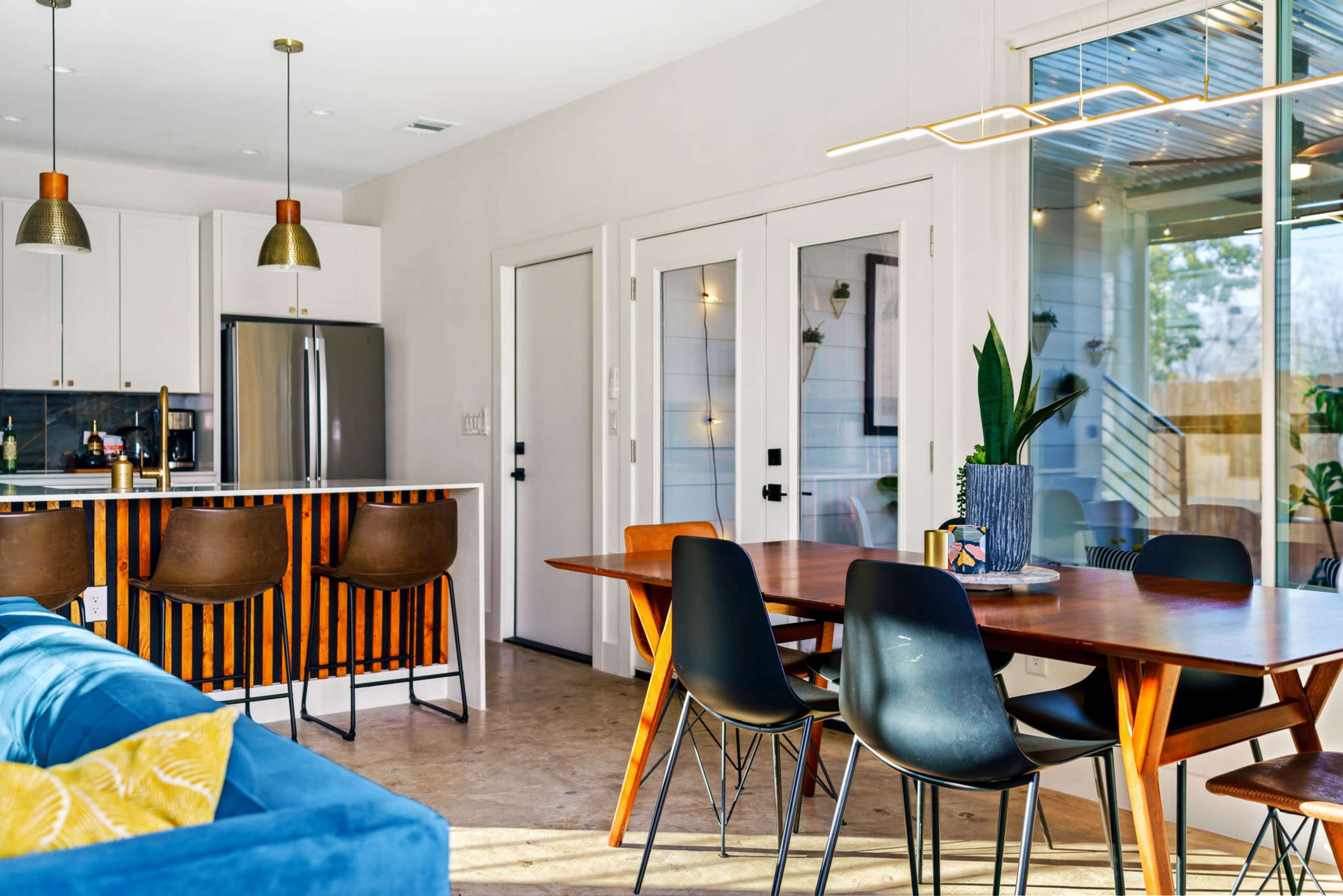 A modern open-concept living space features a kitchen with bar seating, a dining area with a wooden table and black chairs, and large glass doors leading outside.