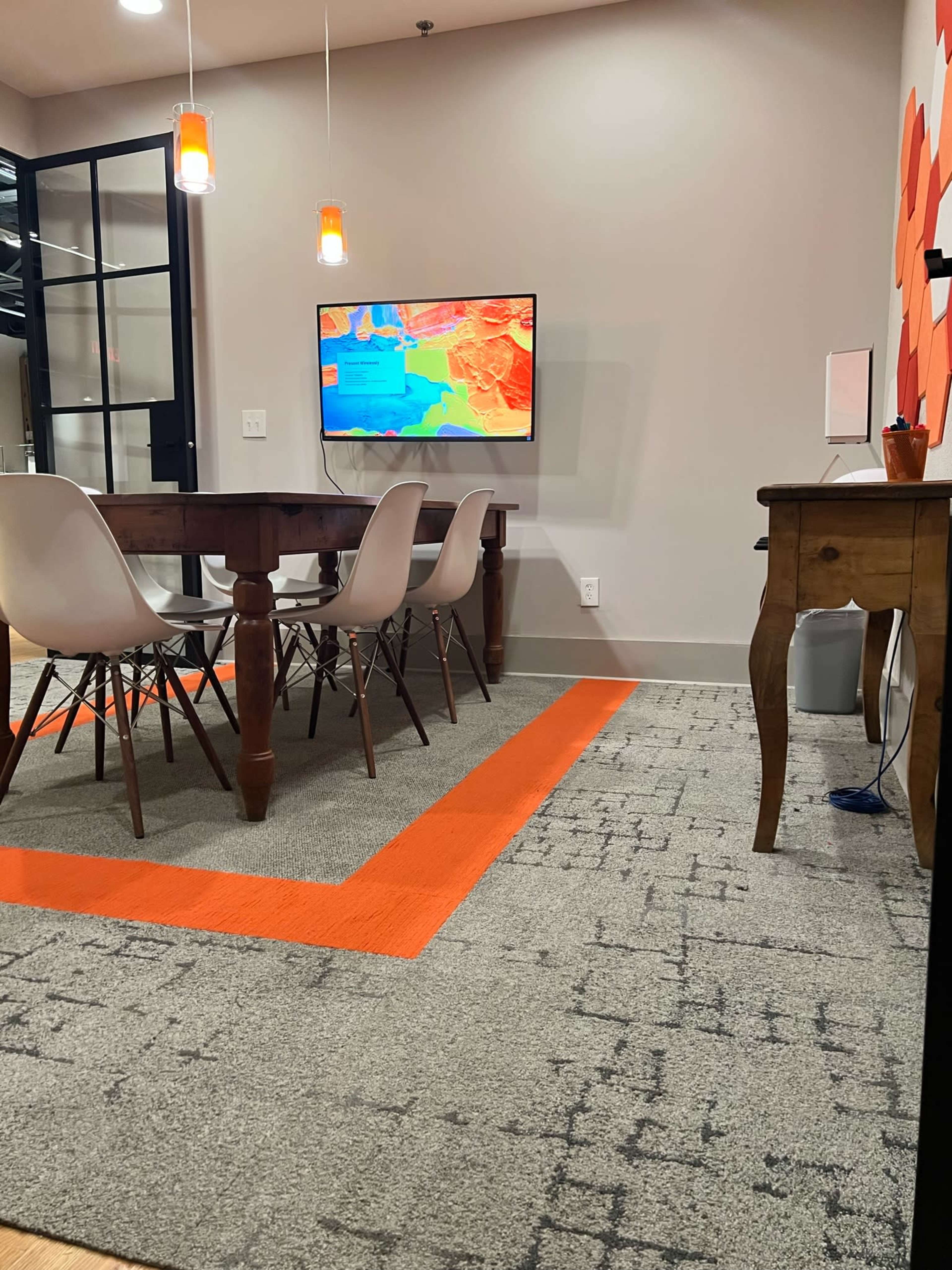 A conference room features a wooden table with several chairs, a wall-mounted TV displaying a colorful map, and an orange accent on the carpet.