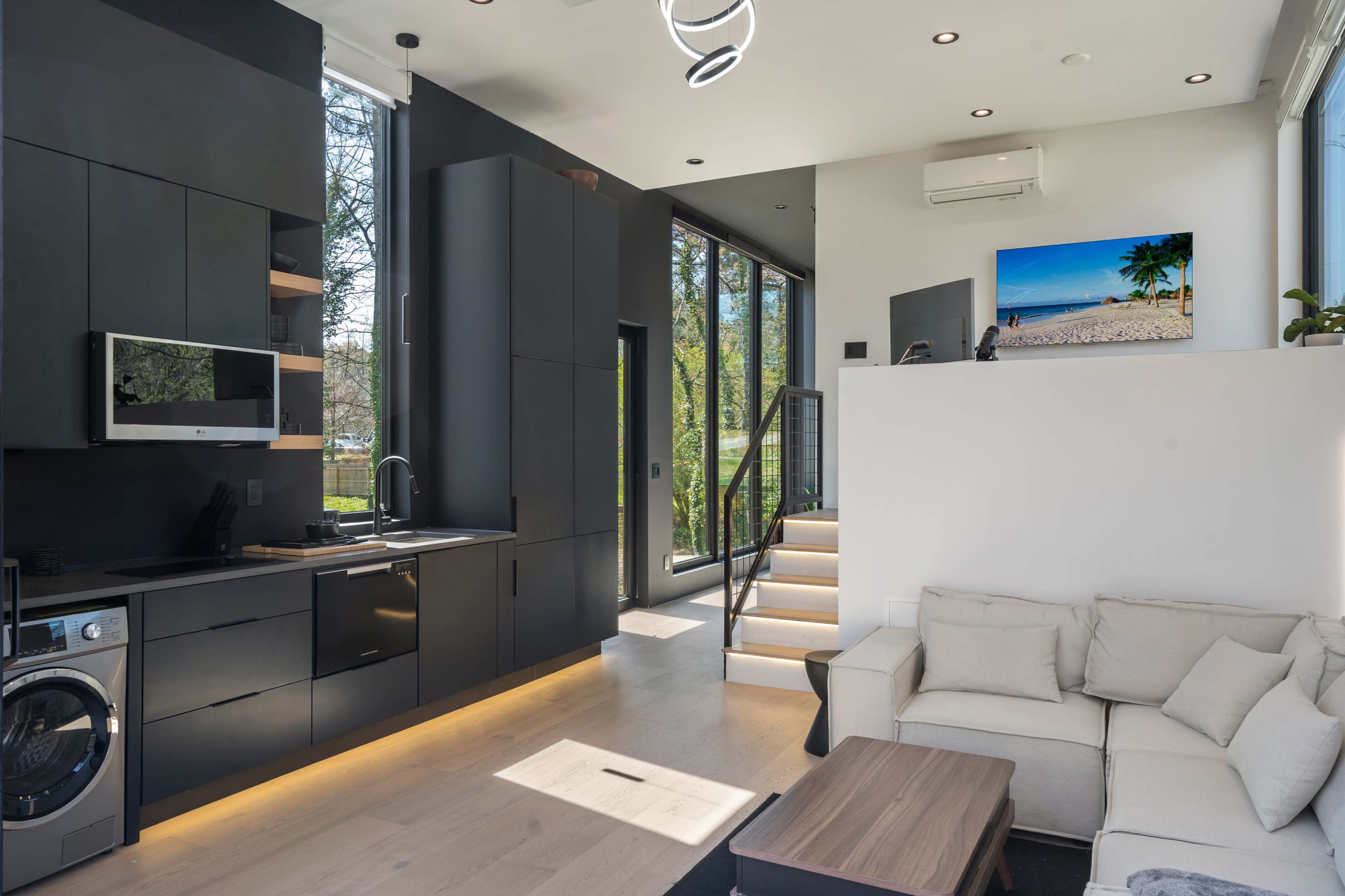 A modern living space featuring a black kitchen, a light-toned sofa, and large windows allowing natural light to fill the room.