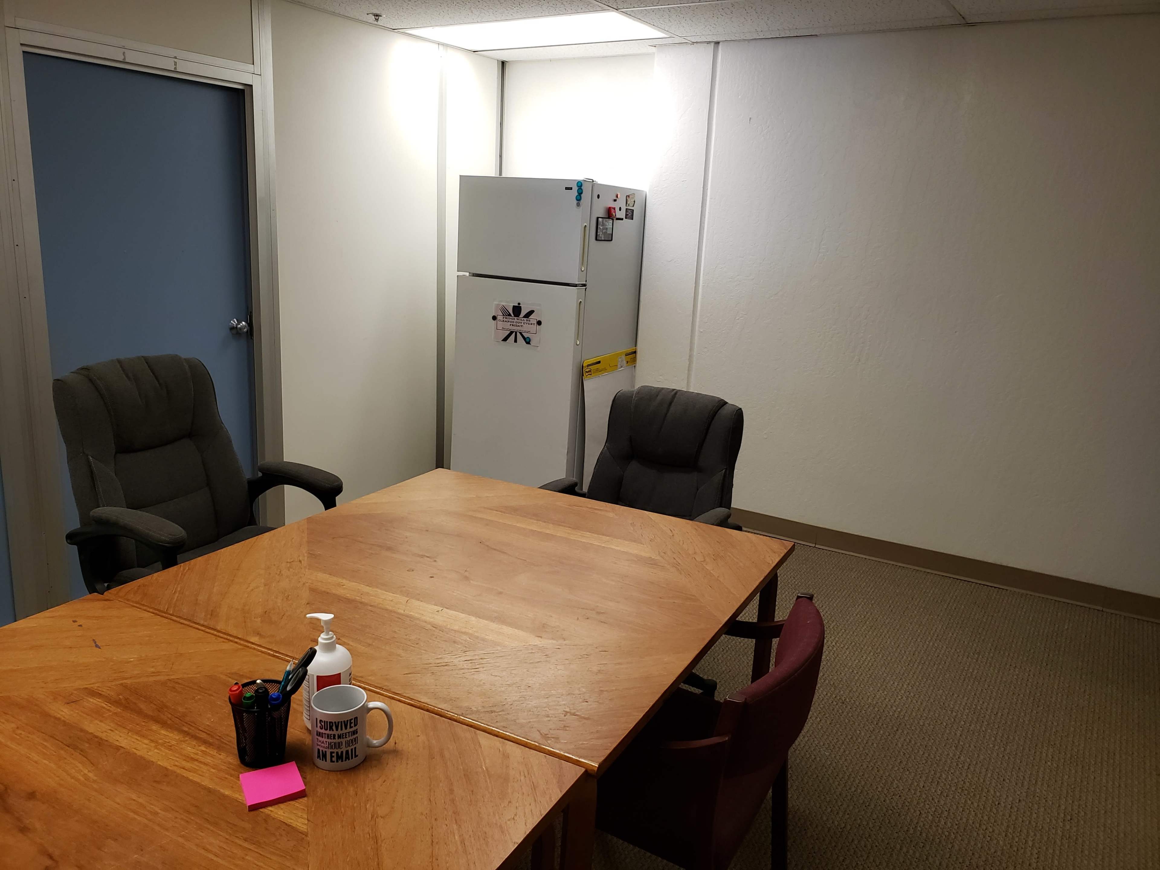 A small empty meeting room features a wooden table, two office chairs, a refrigerator, and a shelf with supplies.
