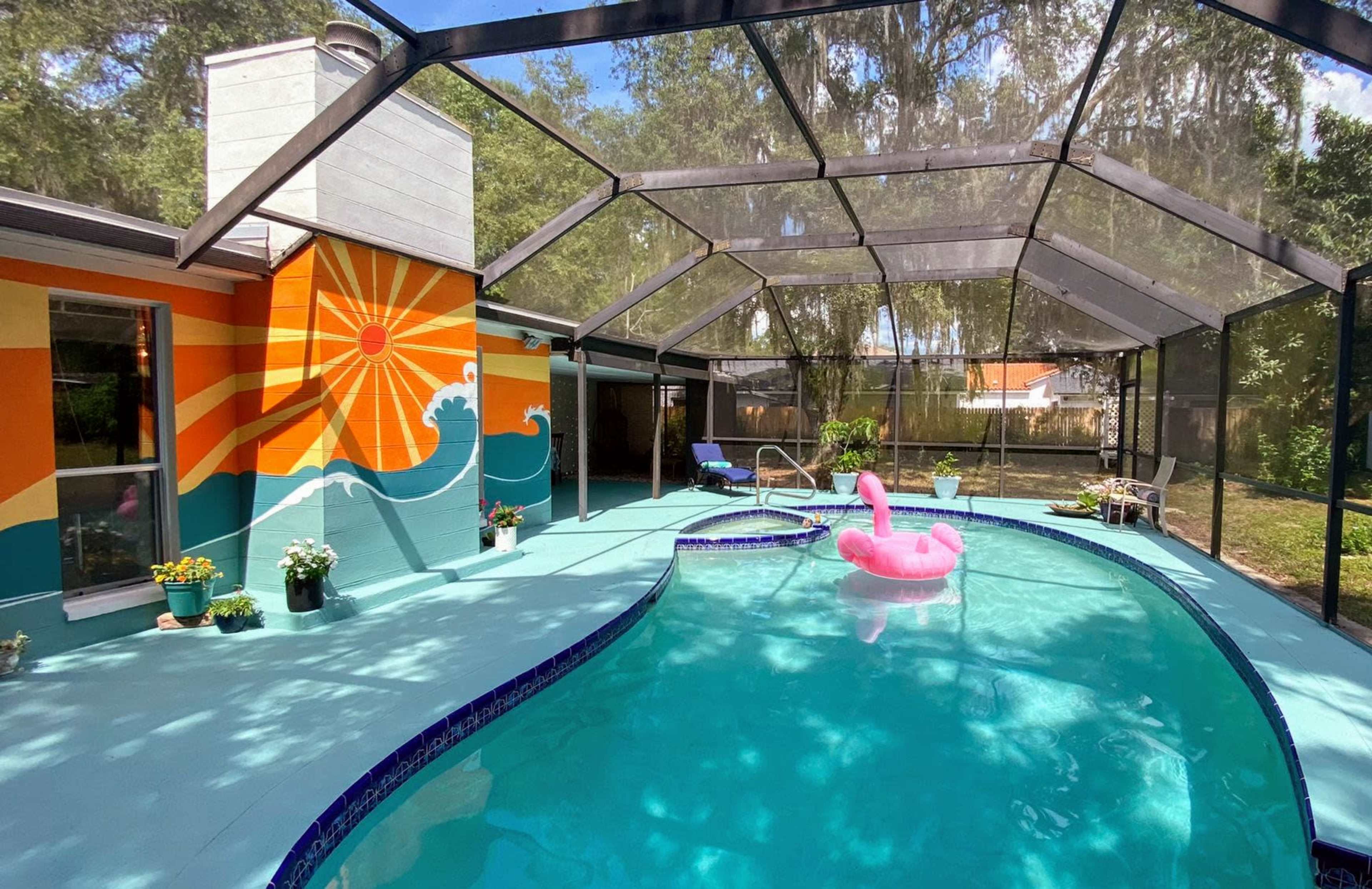 A screened-in pool area with a colorful mural featuring a sun and waves, an inflatable flamingo float, and potted plants around the pool.