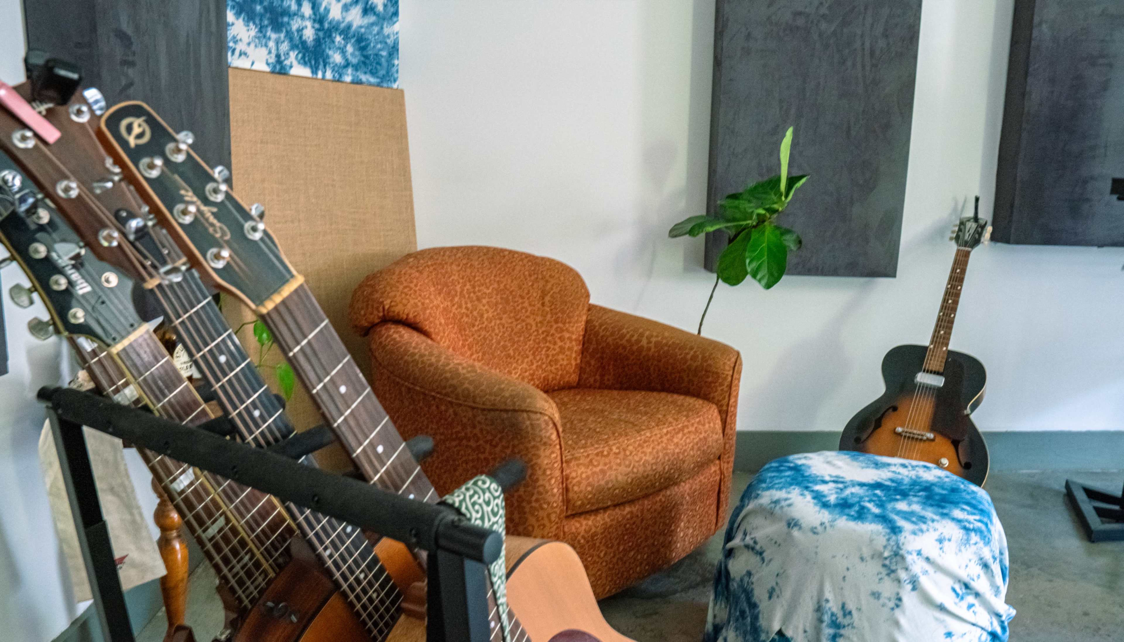 The image shows an indoor music space featuring several guitars on stands, a patterned armchair, a potted plant, and a stool.