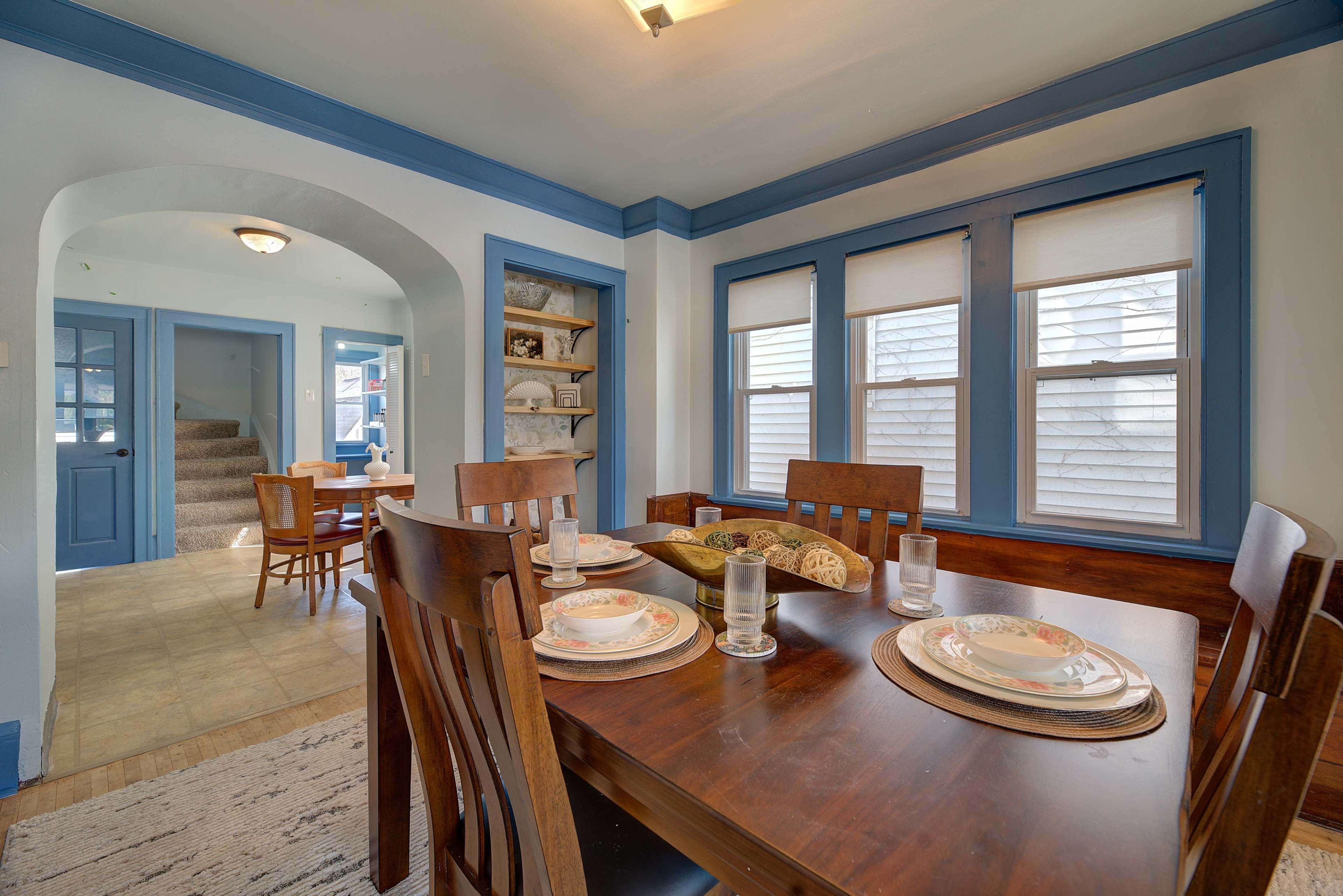 A dining room with a wooden table set for a meal, surrounded by chairs, and features a doorway leading to a stairway and another room.