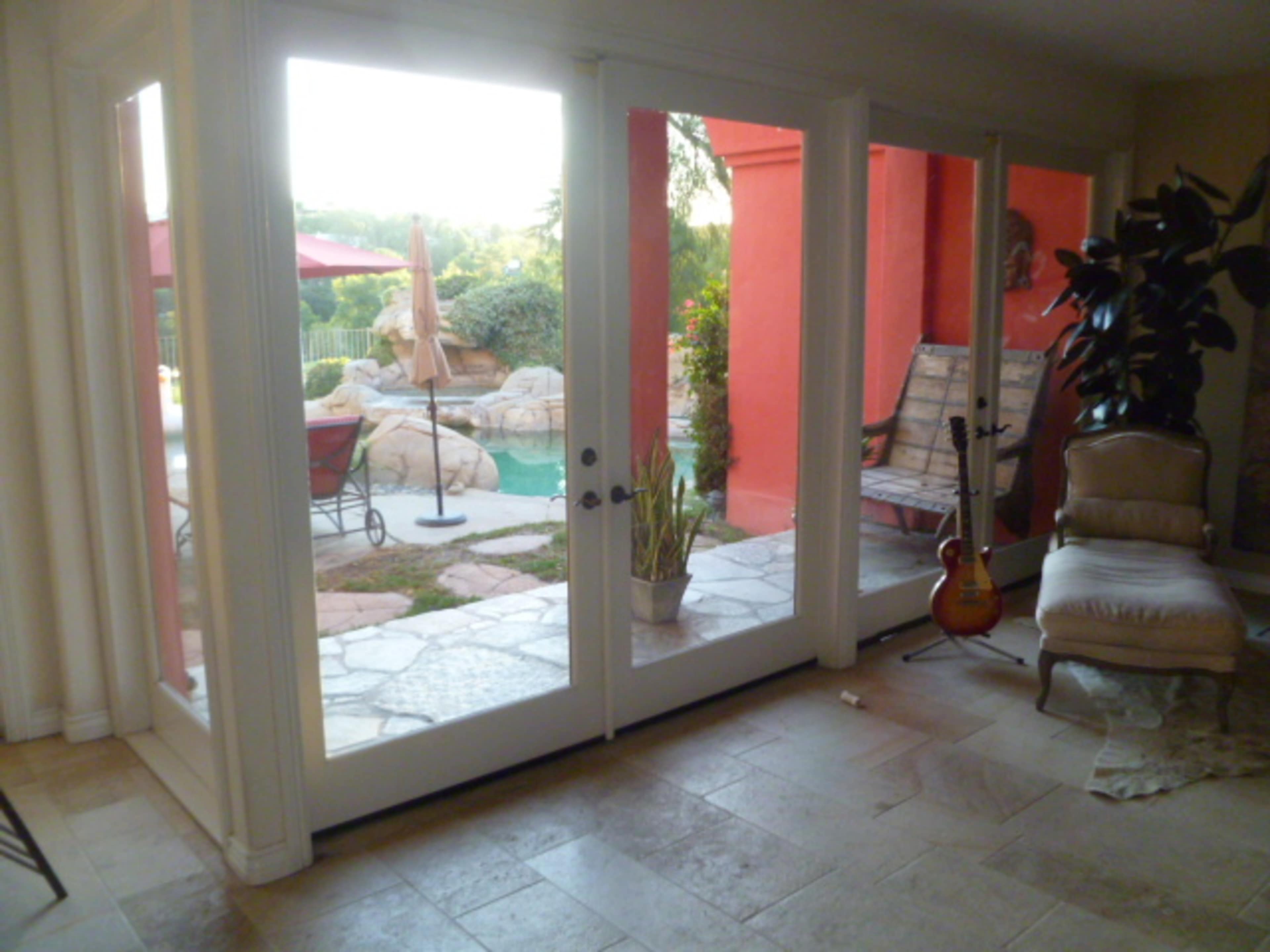 A view through sliding glass doors shows a patio area with a pool, surrounded by rocks and plants, and a red wall in the background.