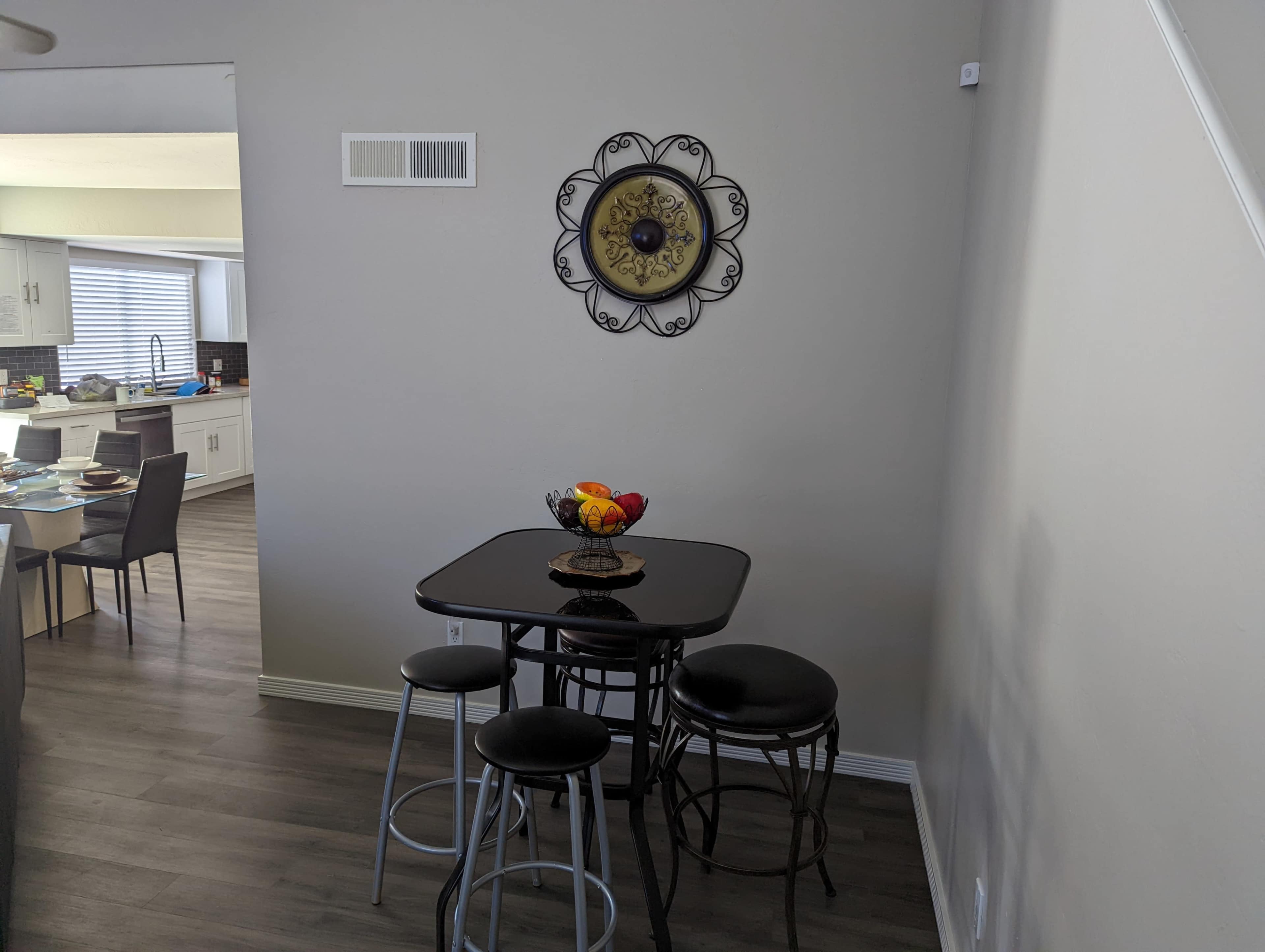 A small dining area features a round black table with four stools and a fruit bowl, situated against a light-colored wall decorated with a circular metal wall clock.
