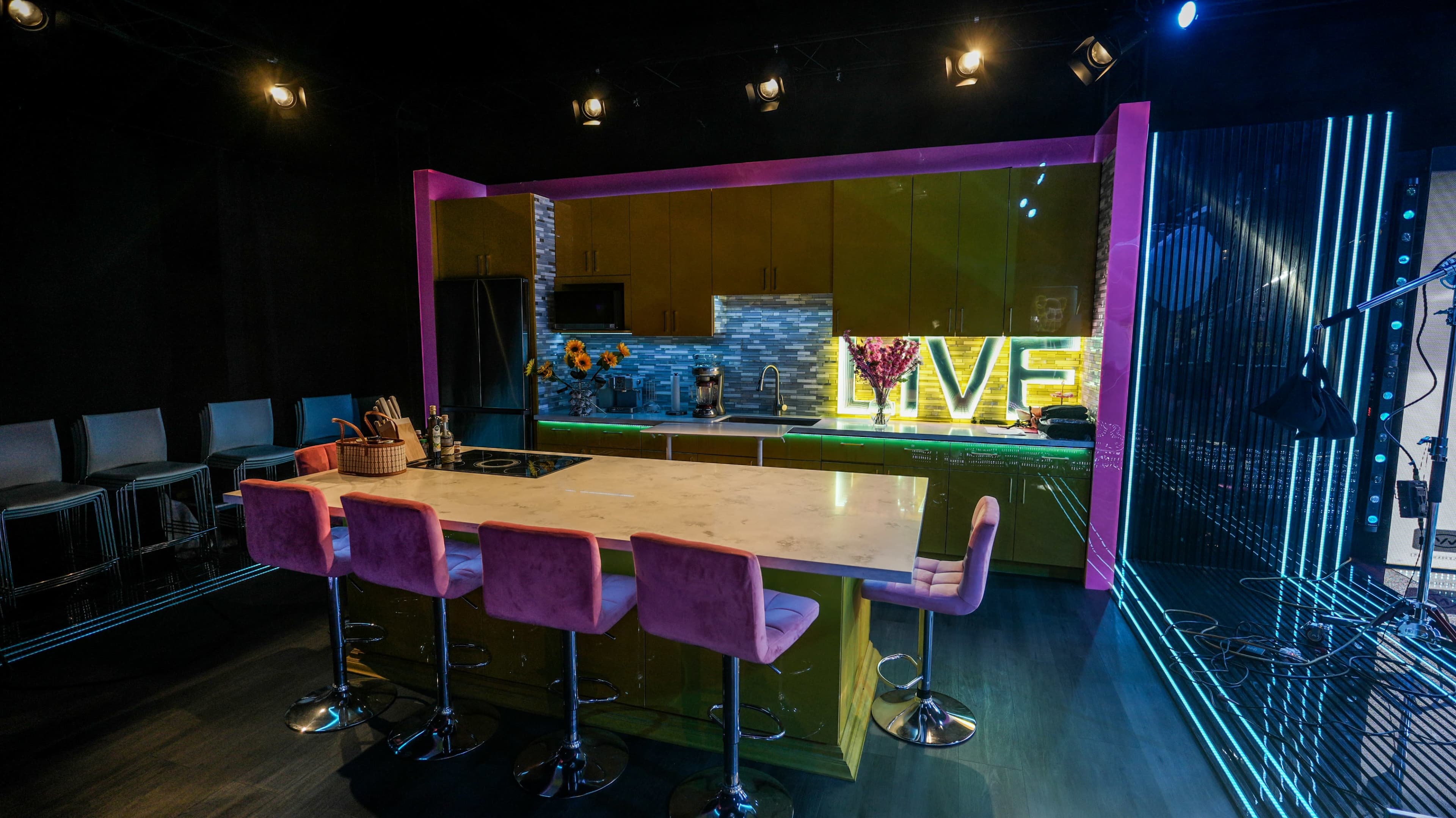 The image shows a modern kitchen studio setting with a long bar counter, colorful cabinetry, and illuminated letters spelling "LIVE" against a backdrop of bright decor.