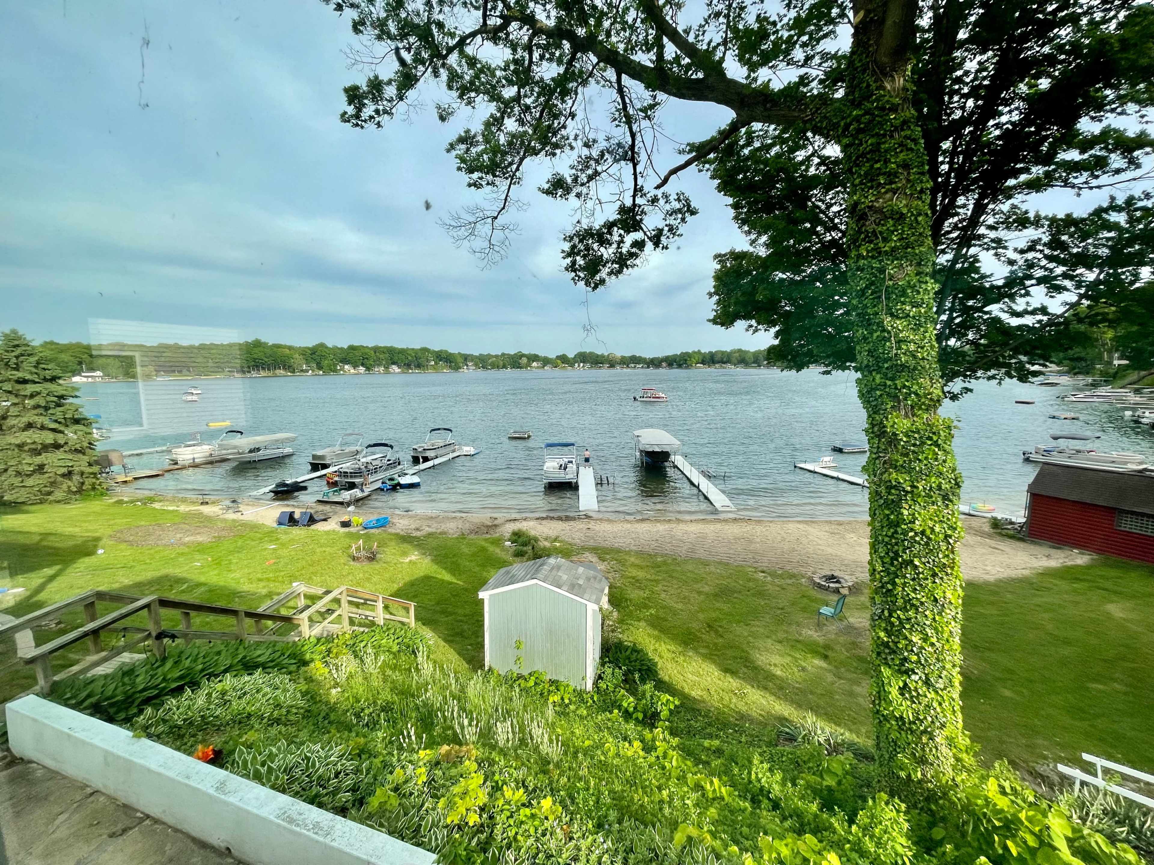 A lakefront scene with several boats docked along the shore and a grassy area with trees and a small shed in the foreground.