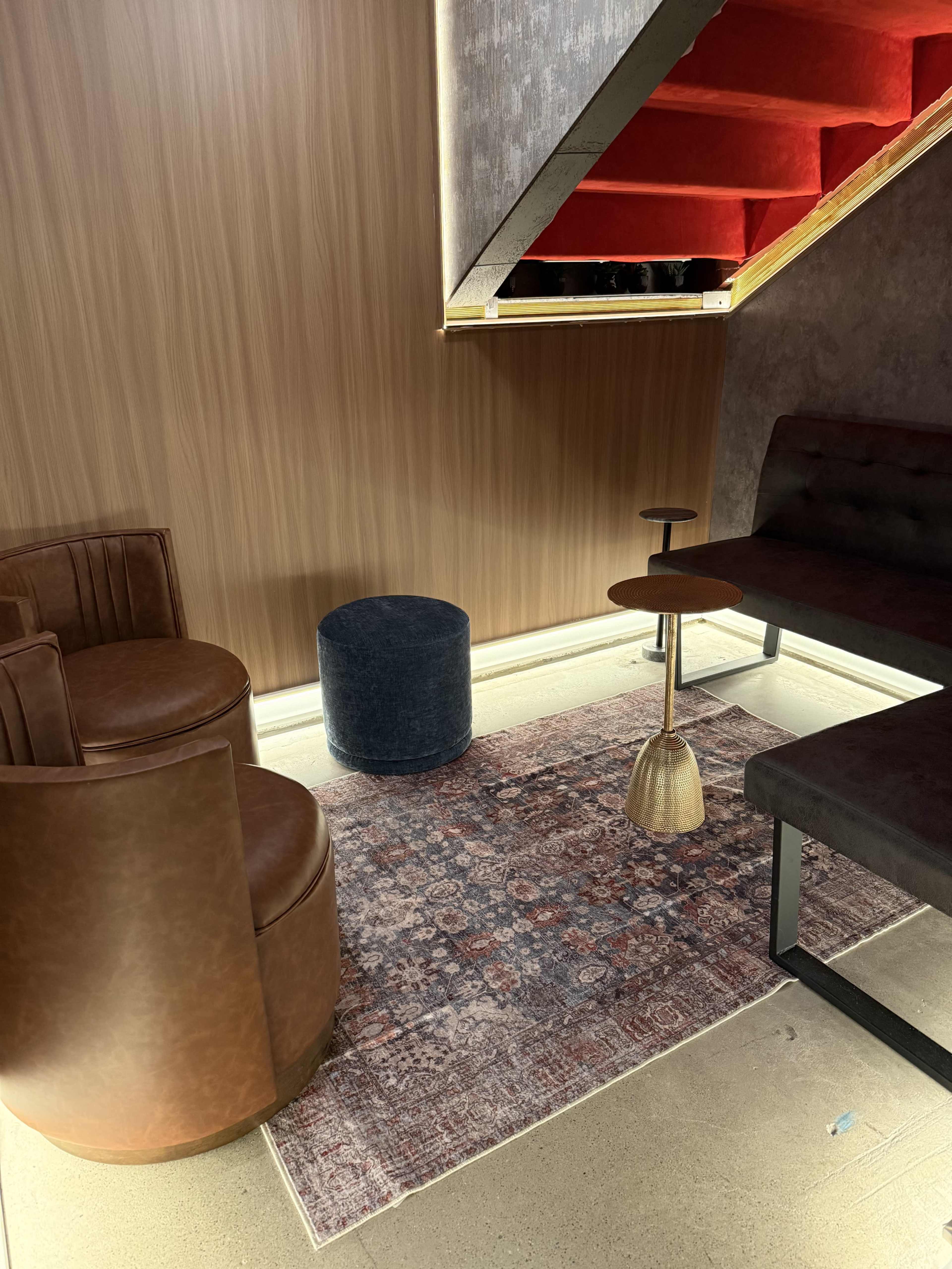 The image shows a cozy seating area beneath a staircase, featuring two brown leather chairs, a round blue ottoman, and a small table on a patterned rug.