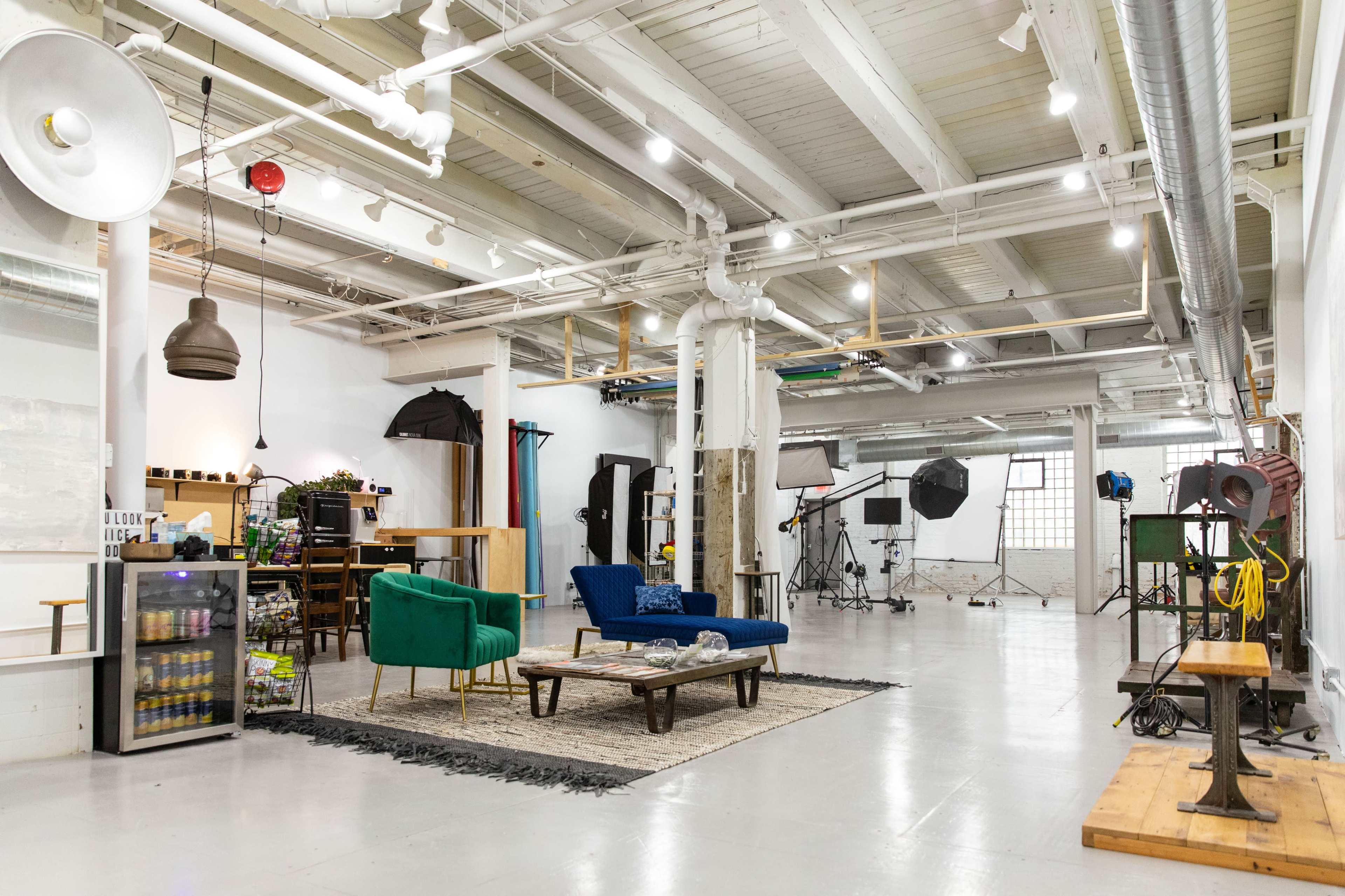 A spacious studio designed for photography, featuring seating areas with blue and green sofas, various lighting equipment, and sets in the background.