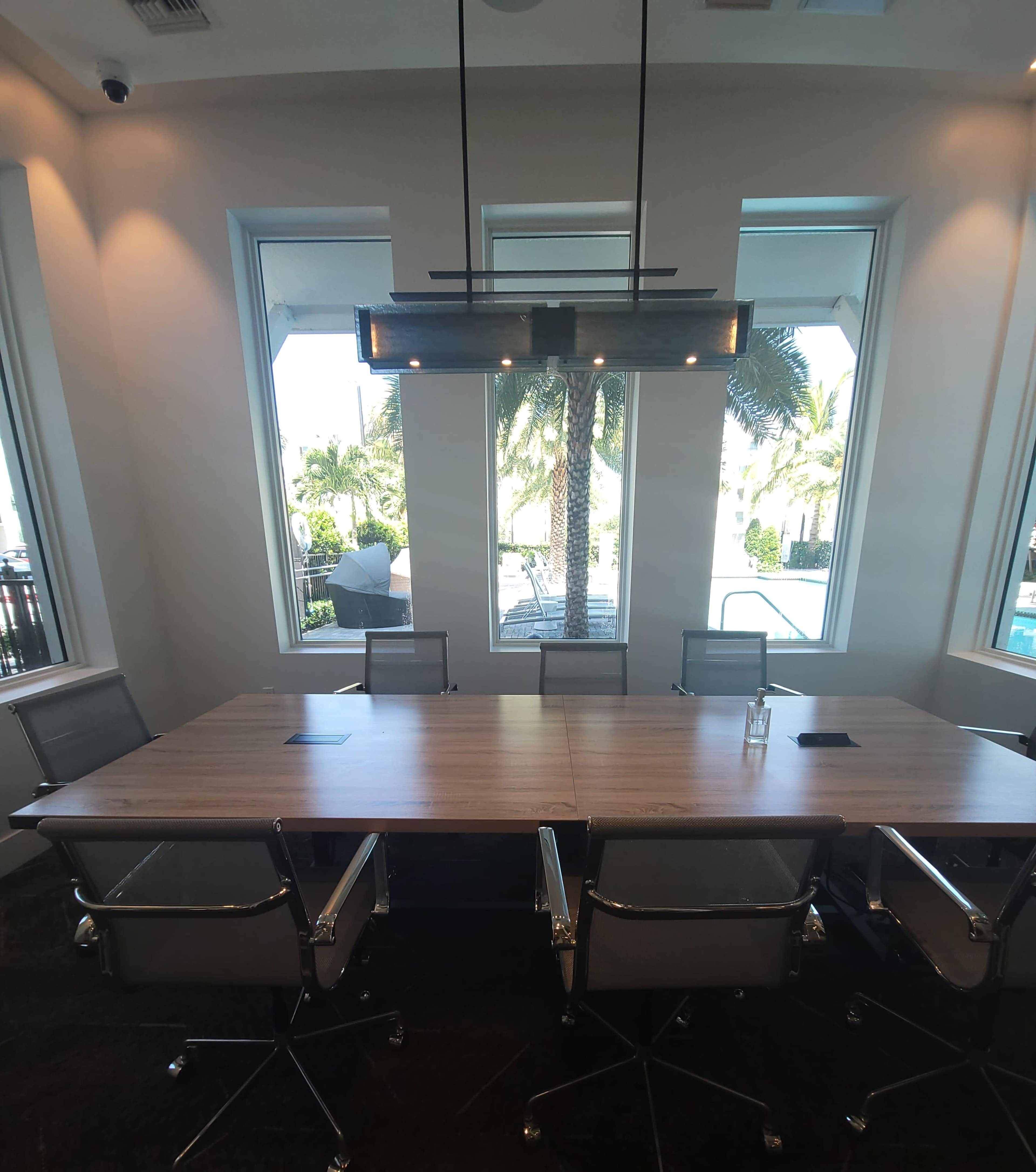 A large, modern conference table with metal-frame chairs is situated in a well-lit meeting room featuring large windows that overlook a pool area and palm trees.