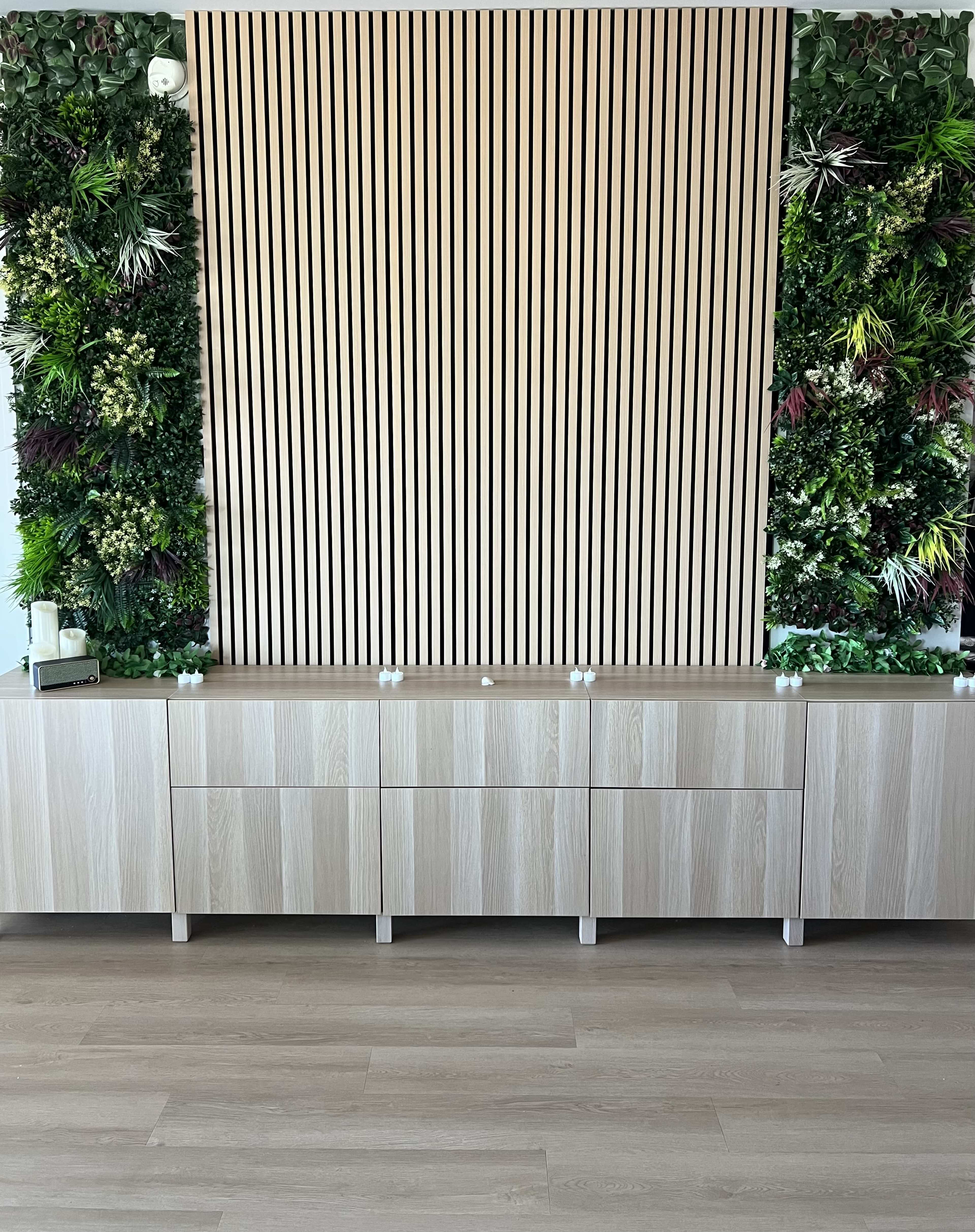 The image shows a contemporary interior space featuring a wooden storage unit below a vertical slatted wood wall, complemented by a lush green vertical garden on either side.