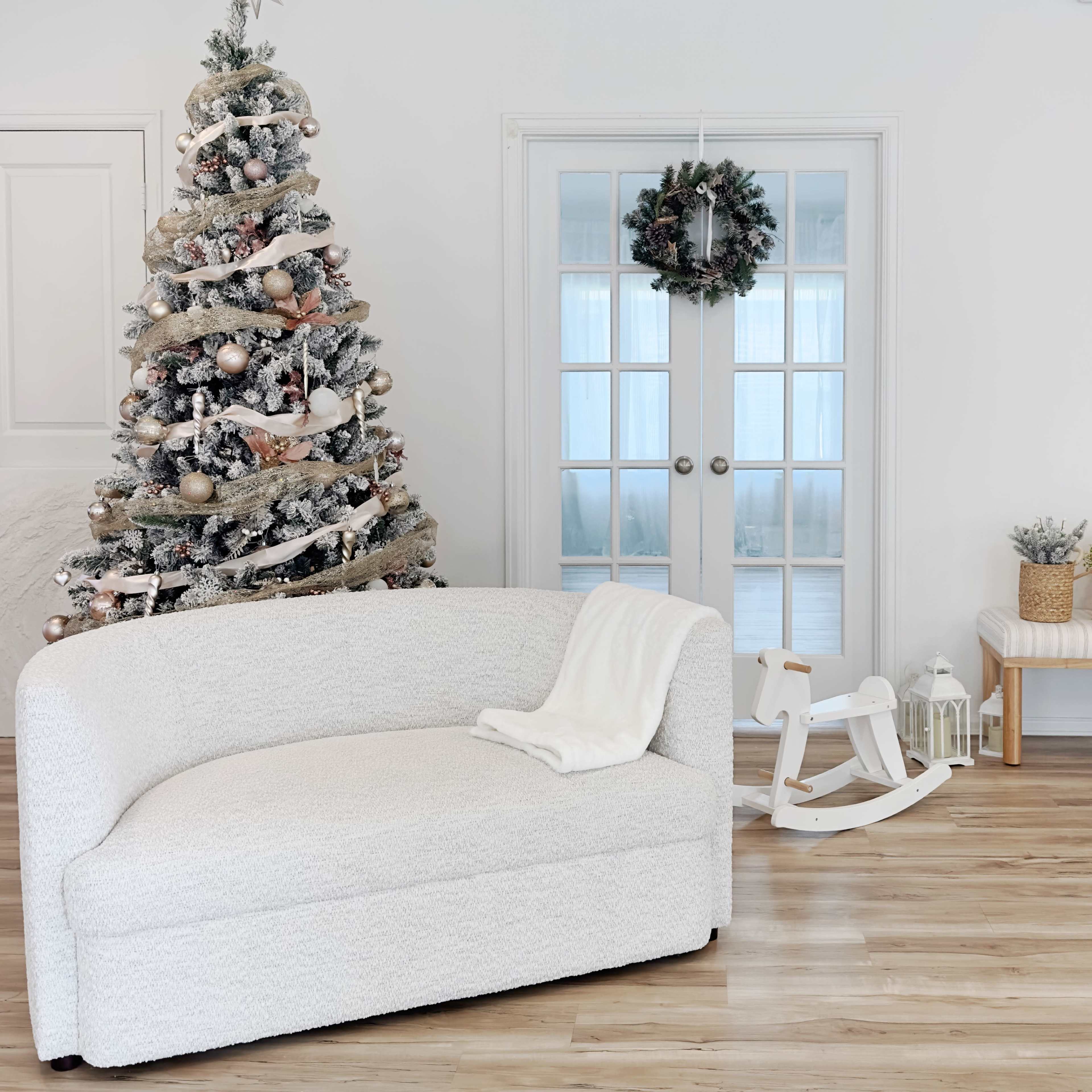 A white curved sofa is positioned near a decorated Christmas tree in a brightly lit room with double glass doors.