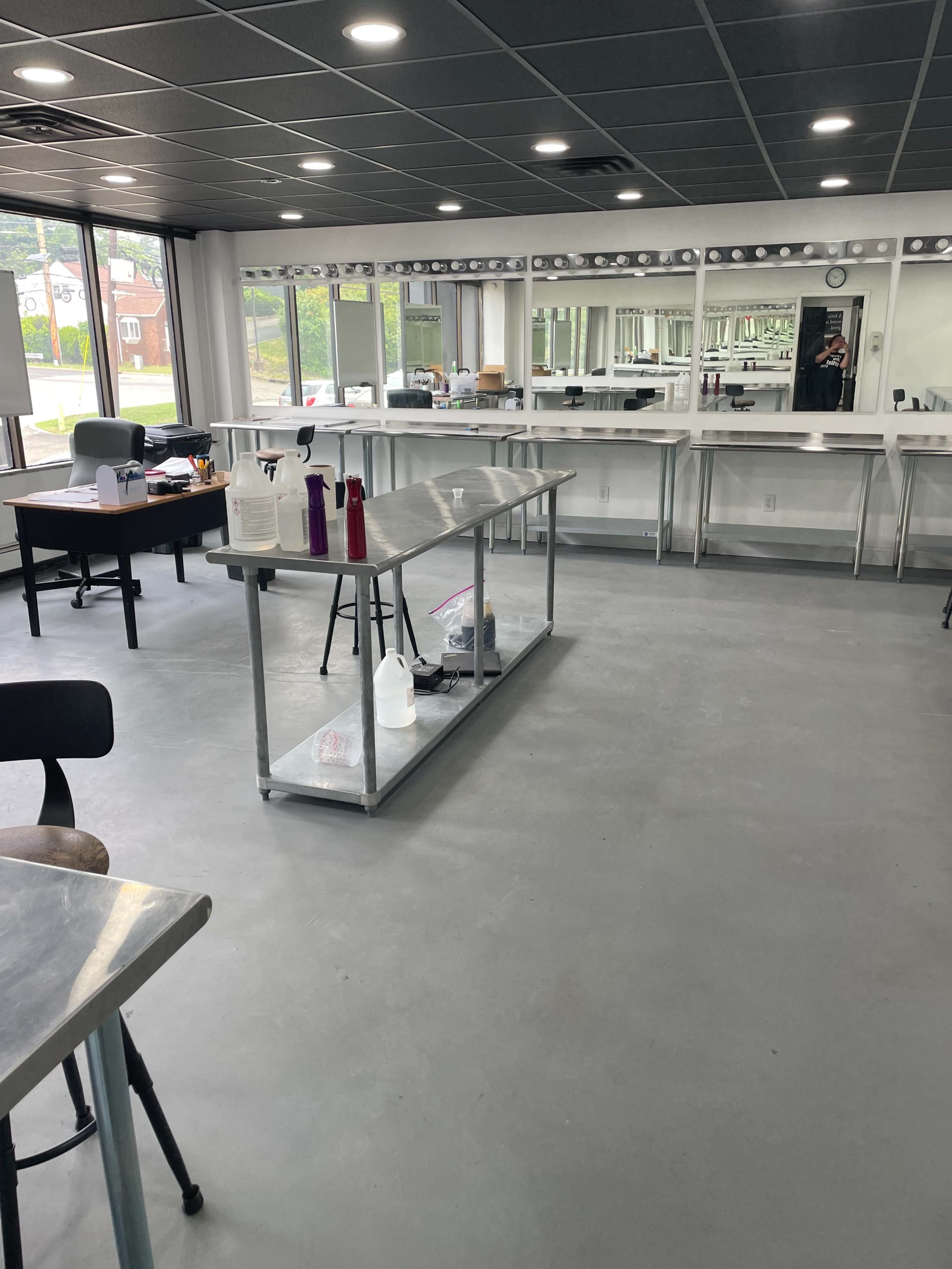 An empty room with a polished concrete floor, large mirrors along one wall, a long table in the center with various bottles, and a desk in the corner.