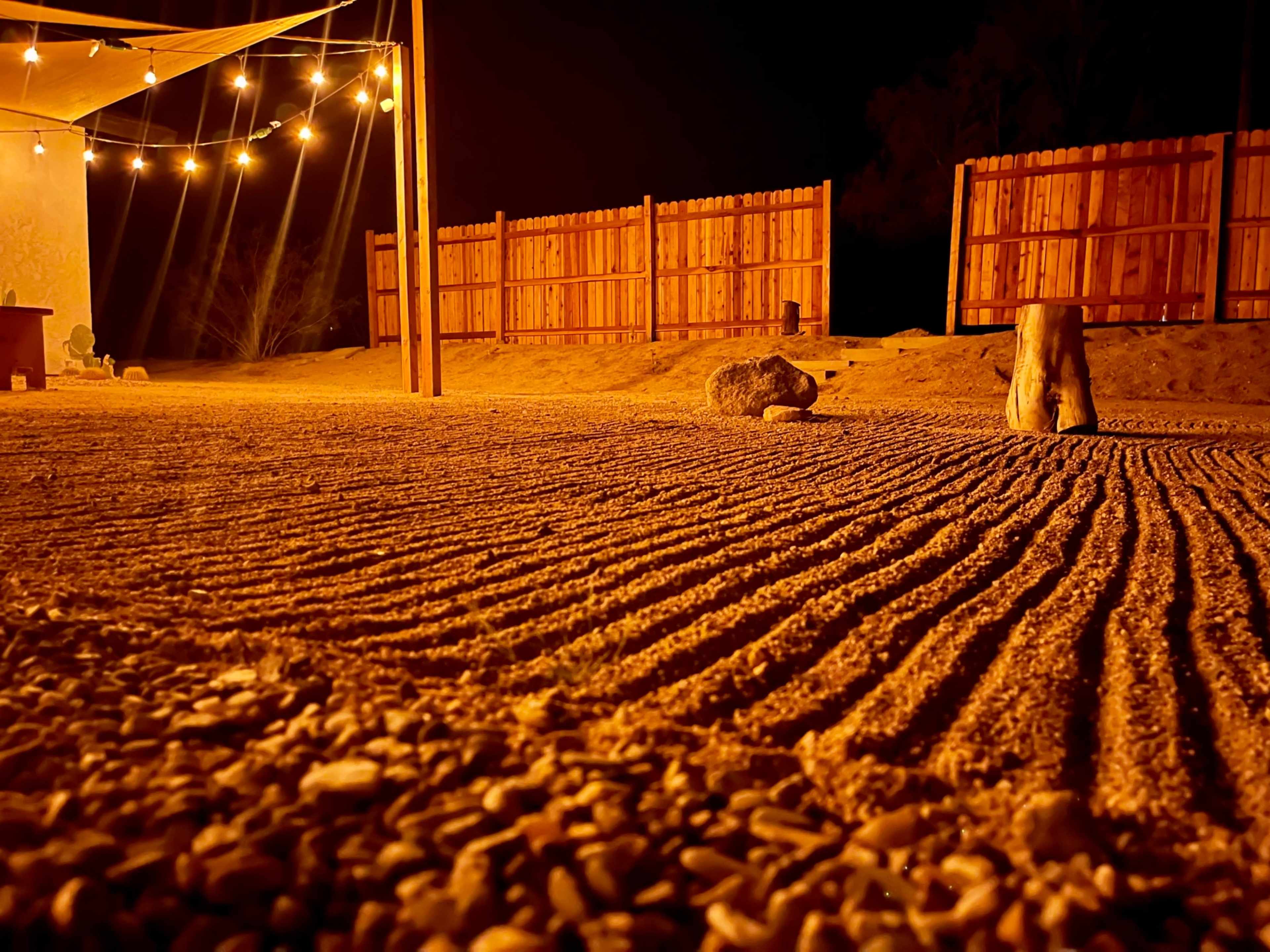 A sandy outdoor area at night with a wooden fence in the background and string lights overhead.