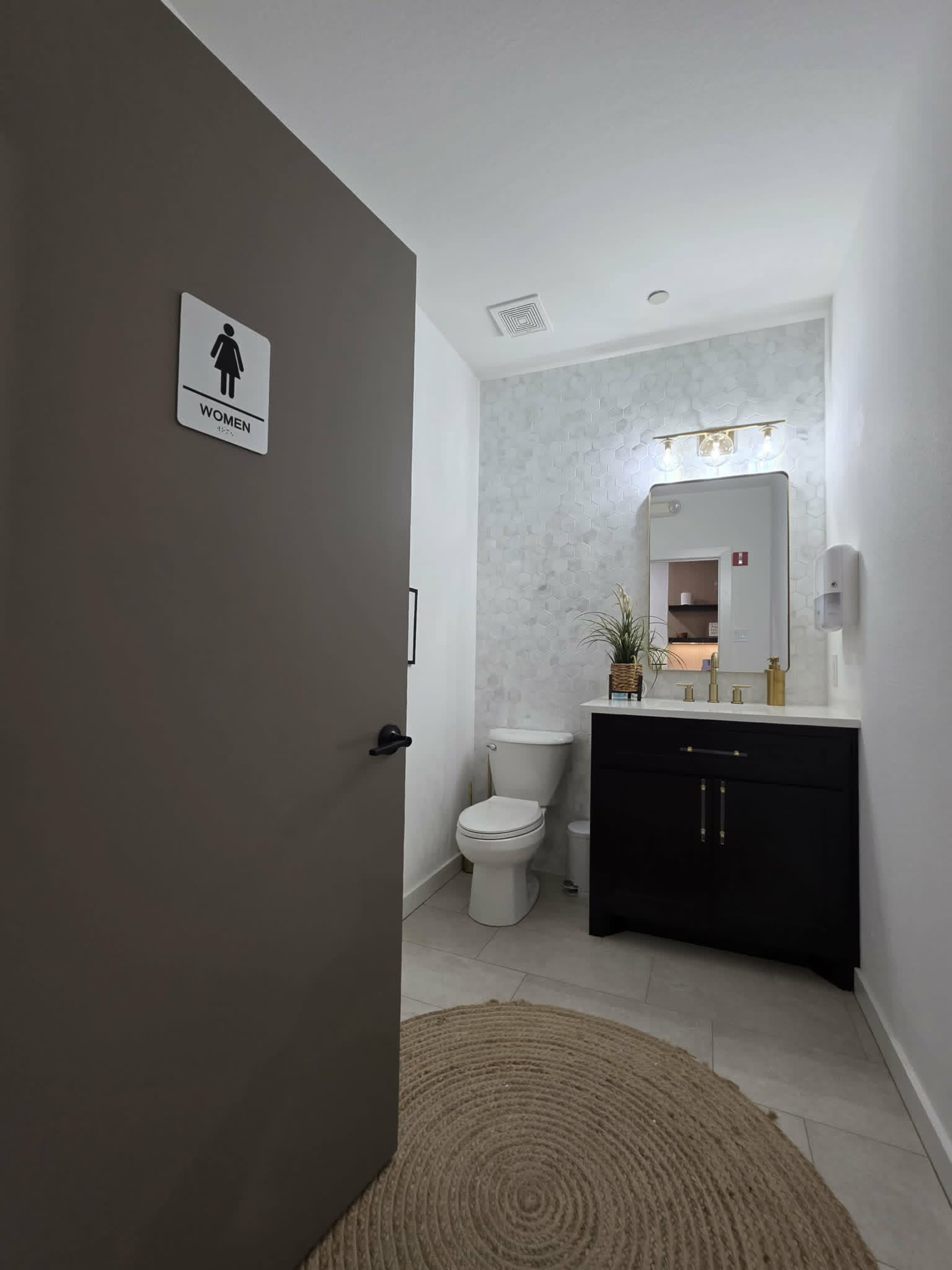 The image shows a women's restroom with a gray door, a white toilet, a wall-mounted mirror, a sink with a dark cabinet, and a circular rug on the tiled floor.
