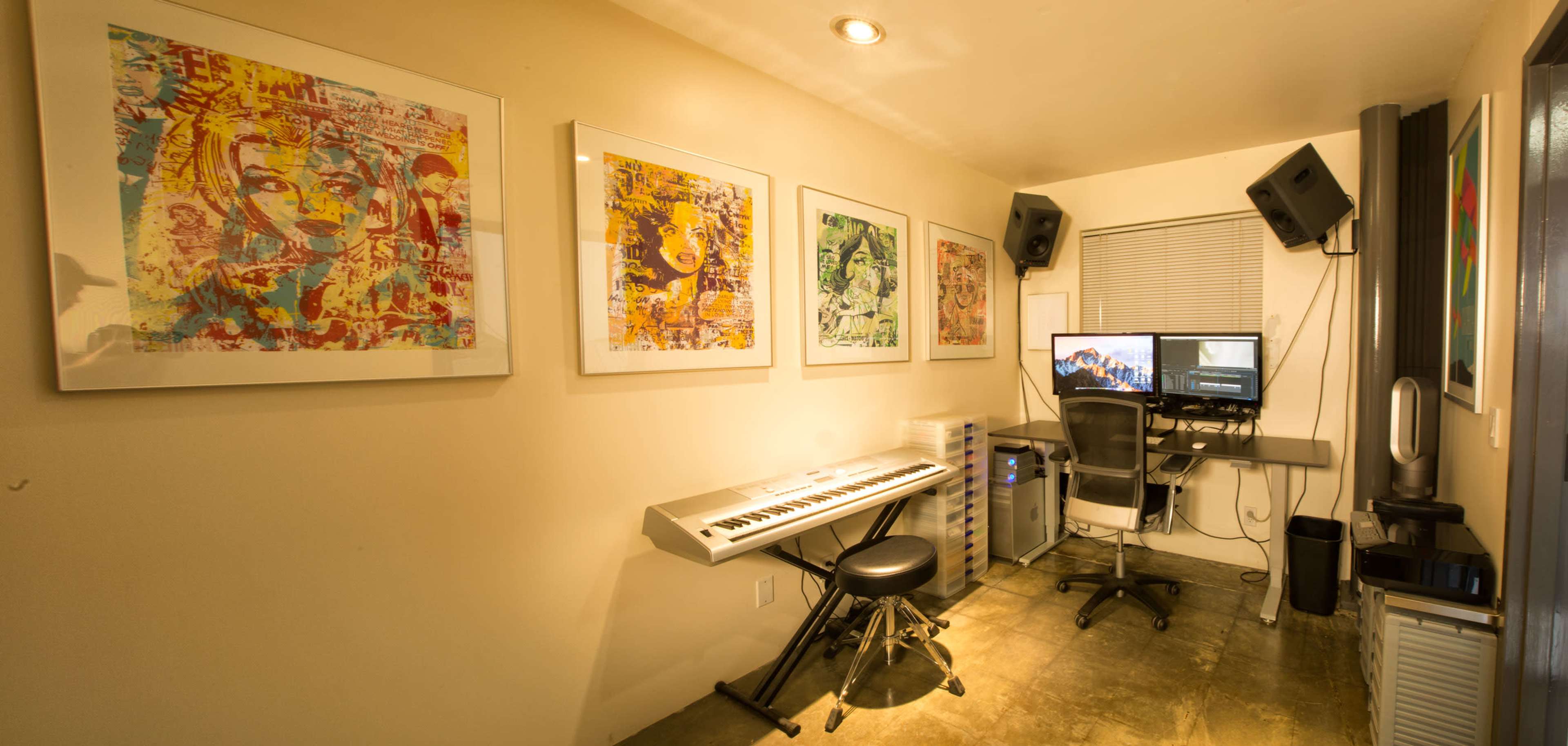 The image shows a music production studio featuring a keyboard, two computer monitors, and artwork on the walls.