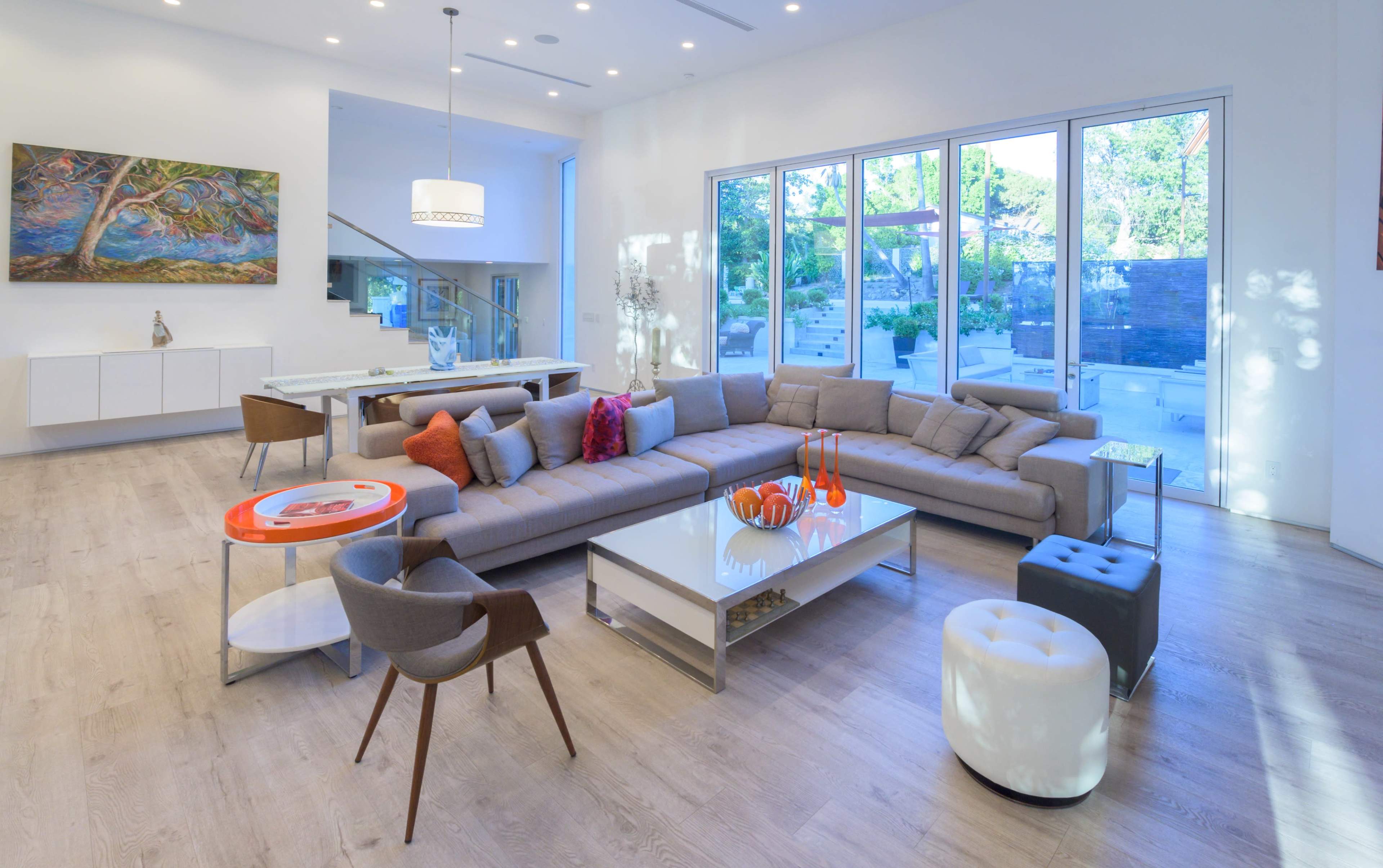 The image shows a modern living room with a large sectional sofa, a glass coffee table, and large windows that overlook an outdoor area.