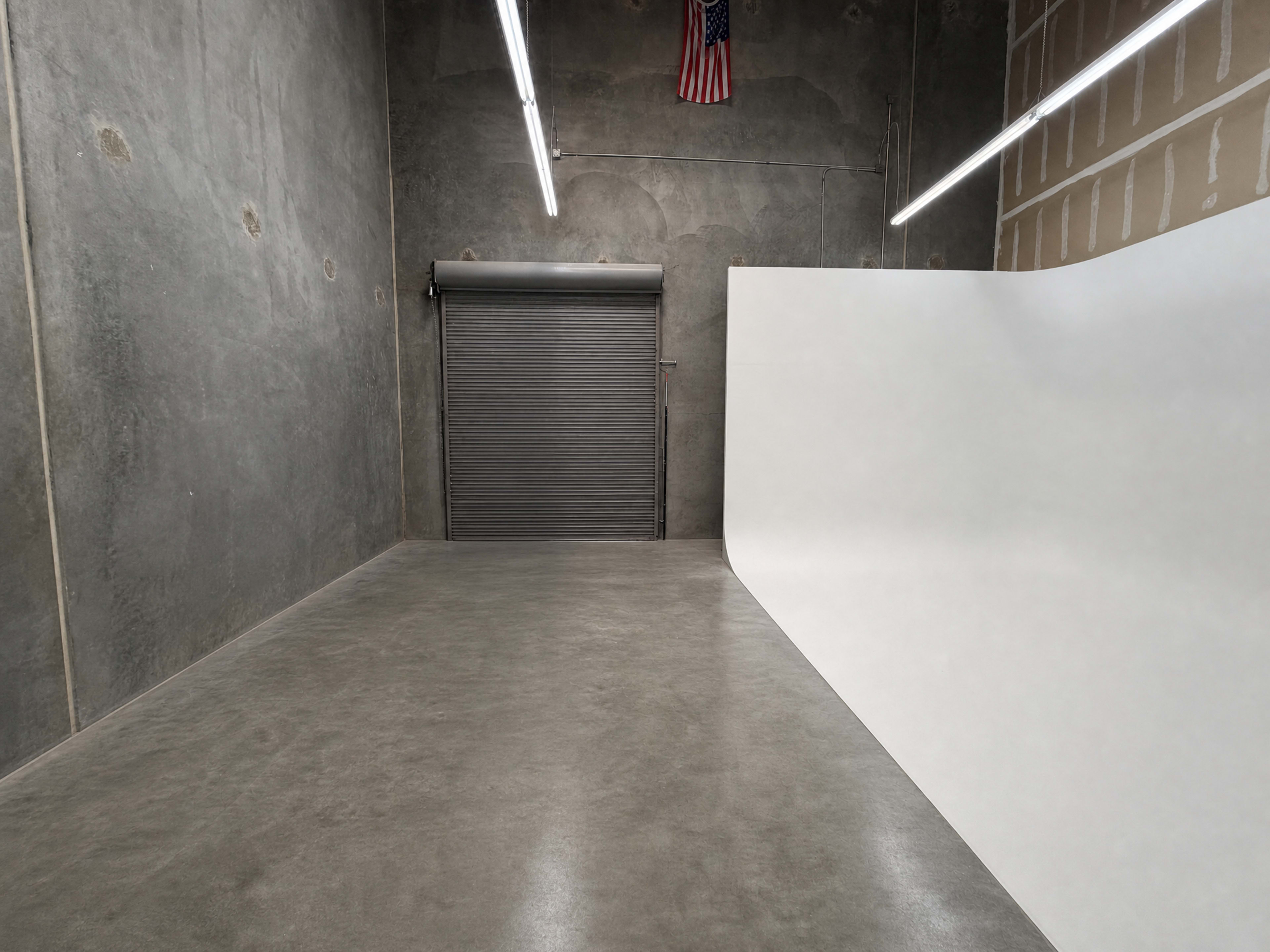 The image shows a spacious, industrial-style room with a concrete floor, a large white backdrop, and a closed roll-up door, along with an American flag hanging on the wall.