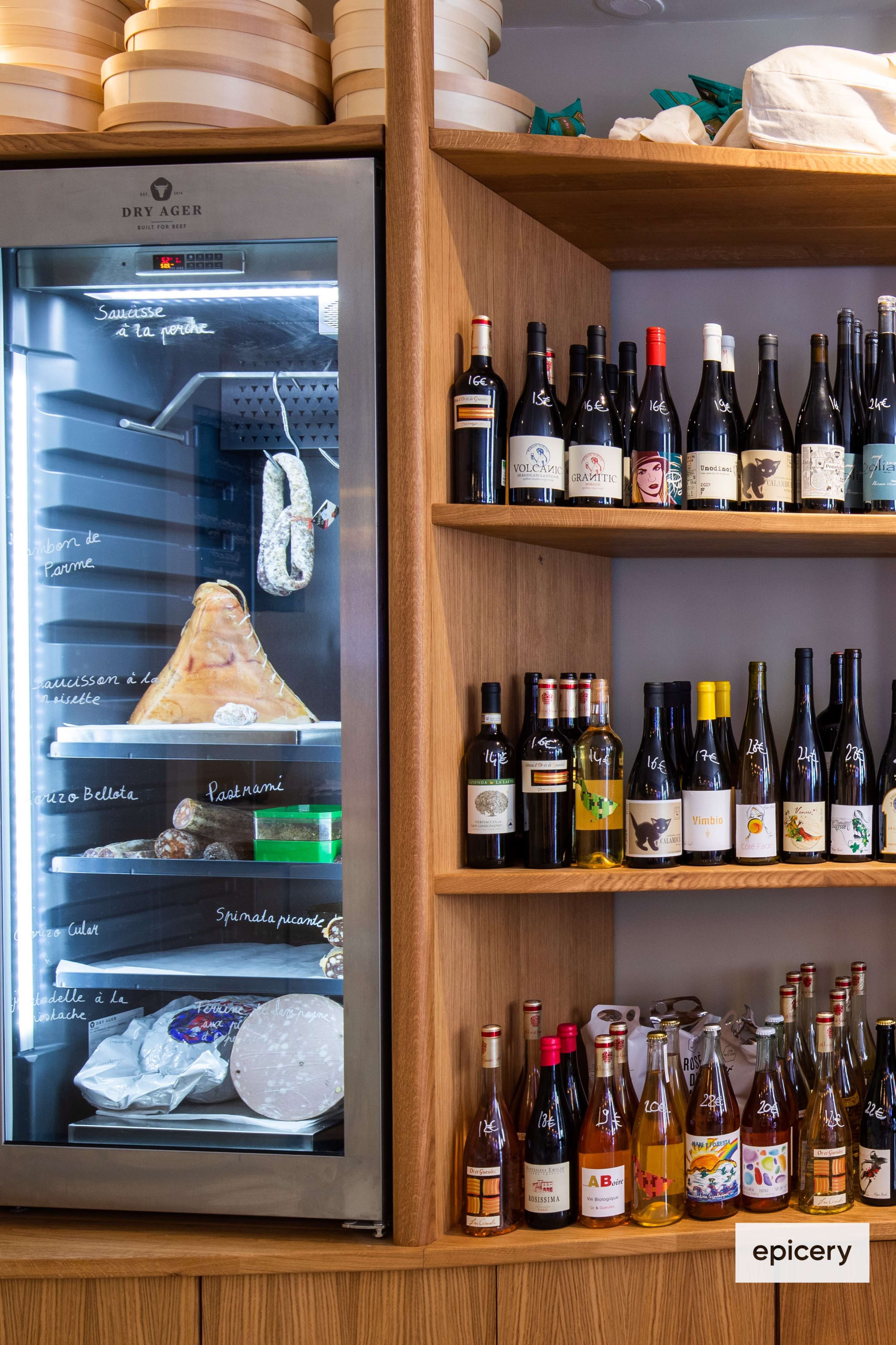 The image shows a wooden shelf displaying various bottles of wine and cider alongside a glass fridge containing a large piece of cheese.