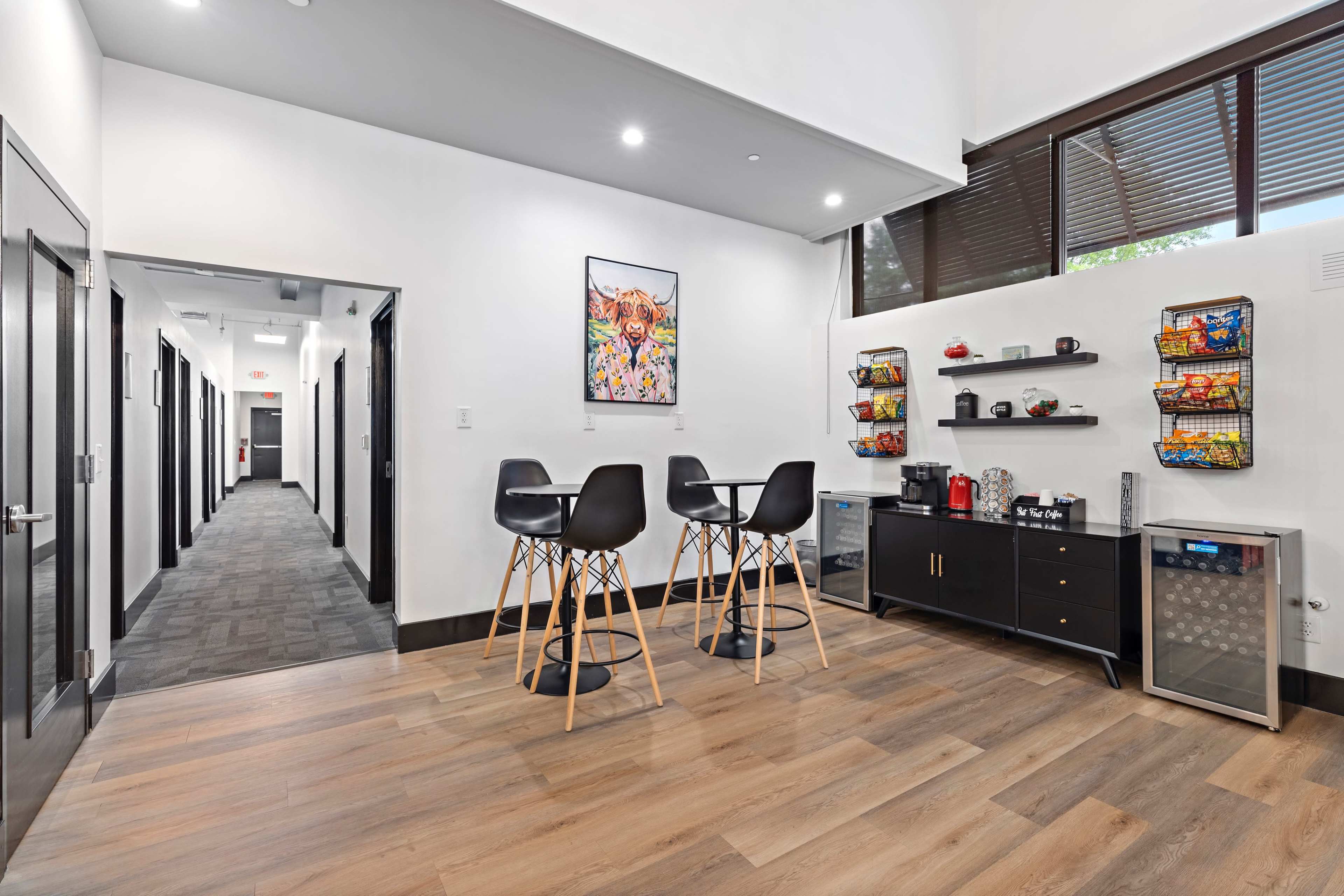 A modern common area with a small dining table and chairs, a kitchenette with snacks, and a corridor leading to multiple closed doors.
