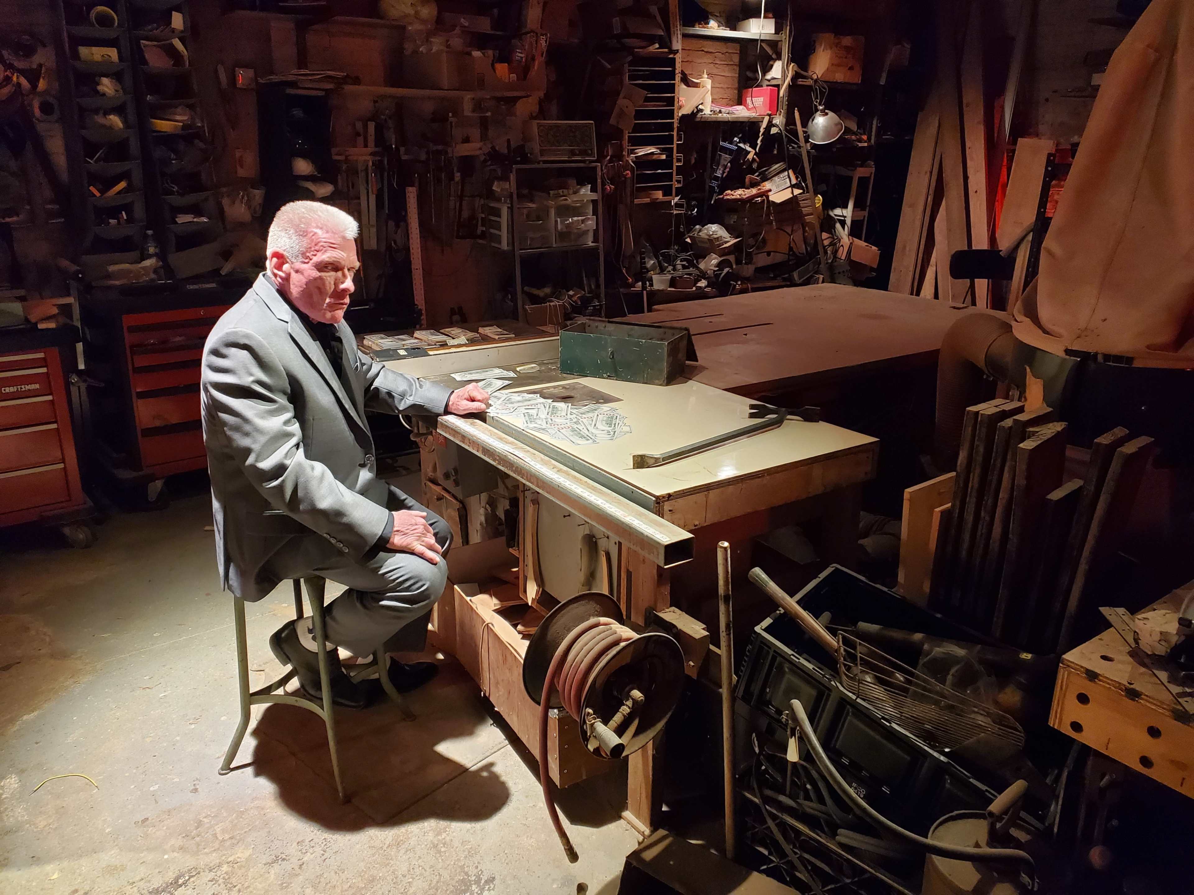 A man in a suit sits on a chair by a woodworking table in a cluttered workshop filled with tools and materials.