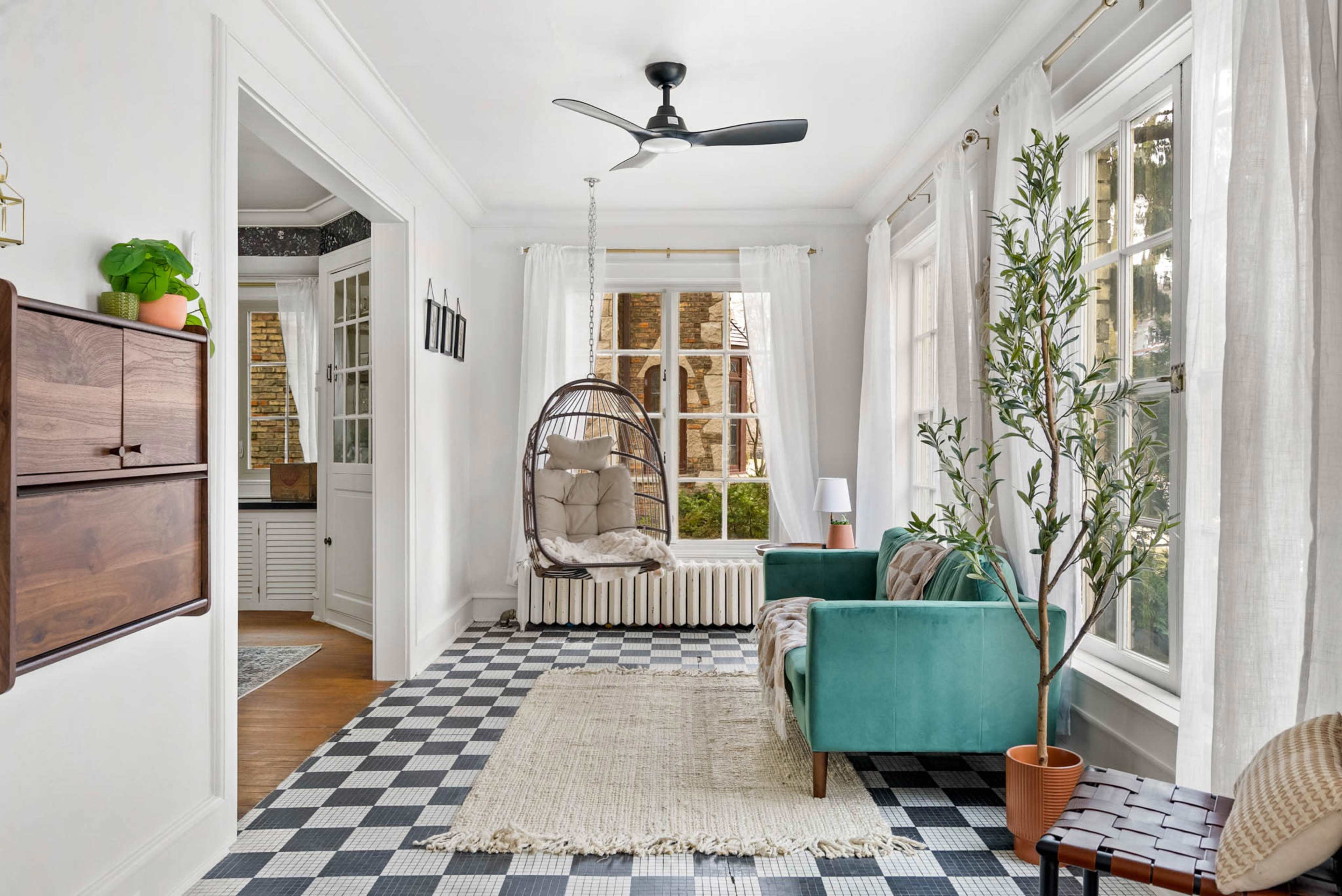 A bright room with a checkered floor, featuring a green sofa, a hanging chair, a small indoor plant, and large windows with white curtains.
