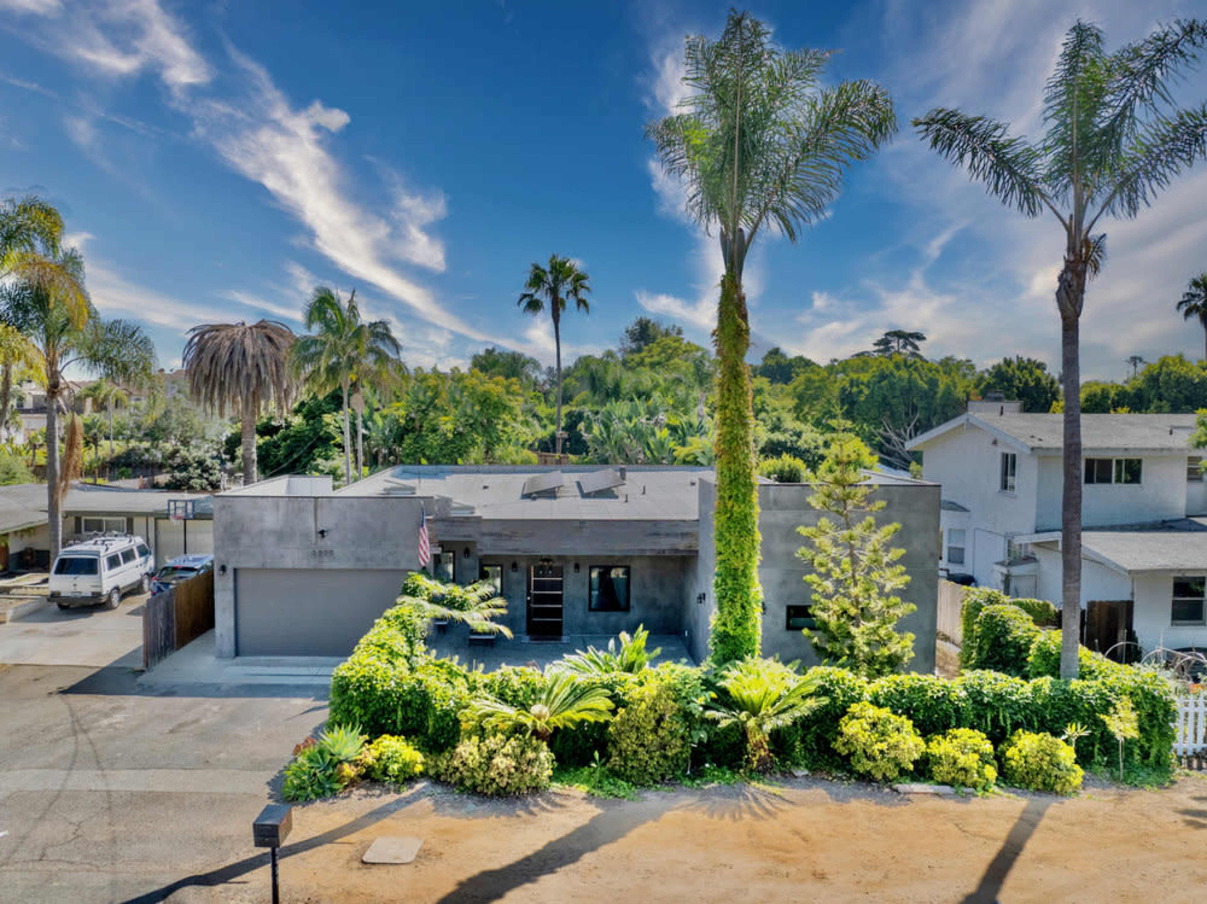 A modern home with palm trees and landscaped greenery is shown in a suburban setting under a blue sky.
