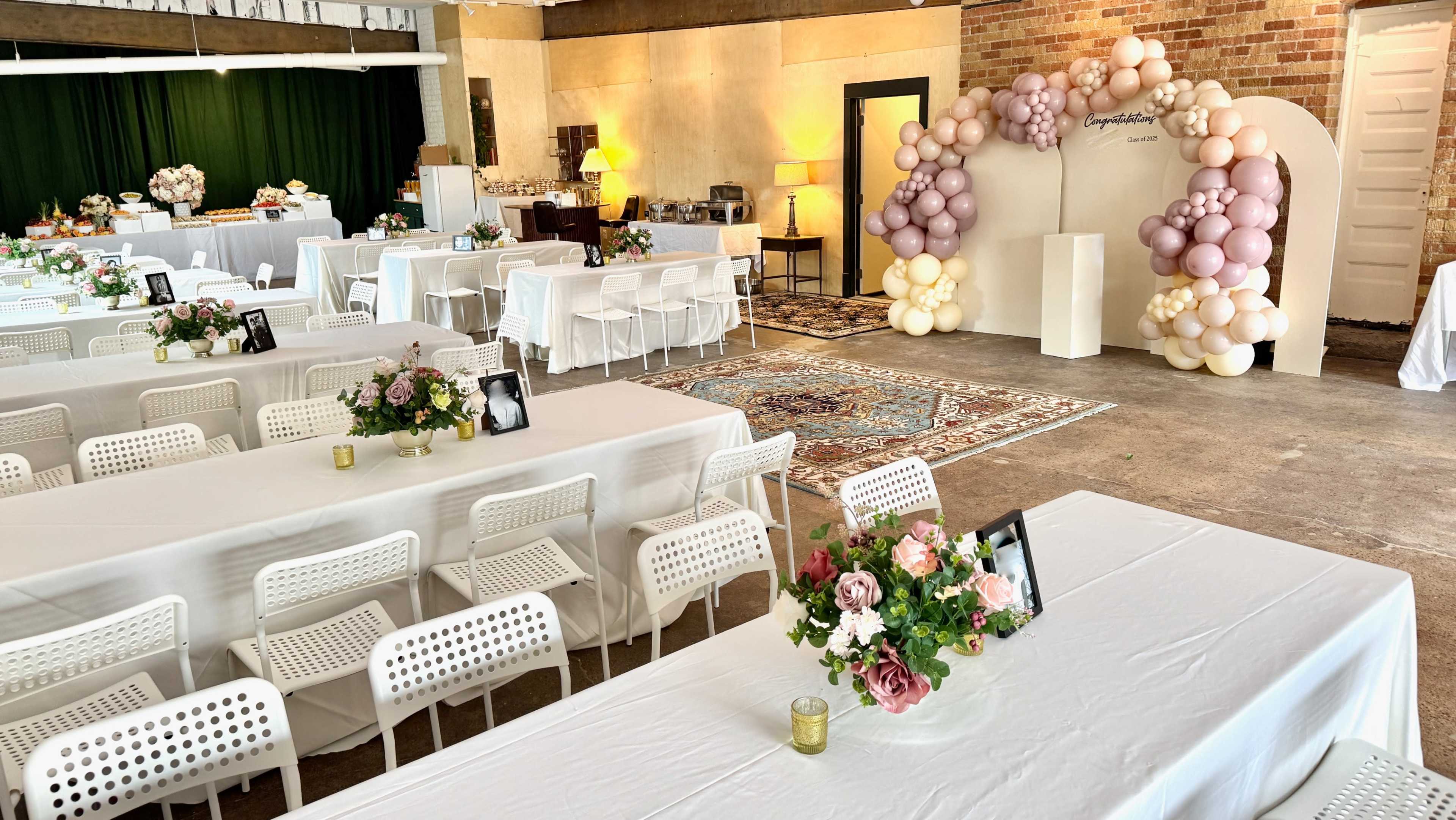 The image shows a decorated indoor space set up for a celebration, featuring tables with floral centerpieces, a balloon arch, and a vintage rug on the floor.