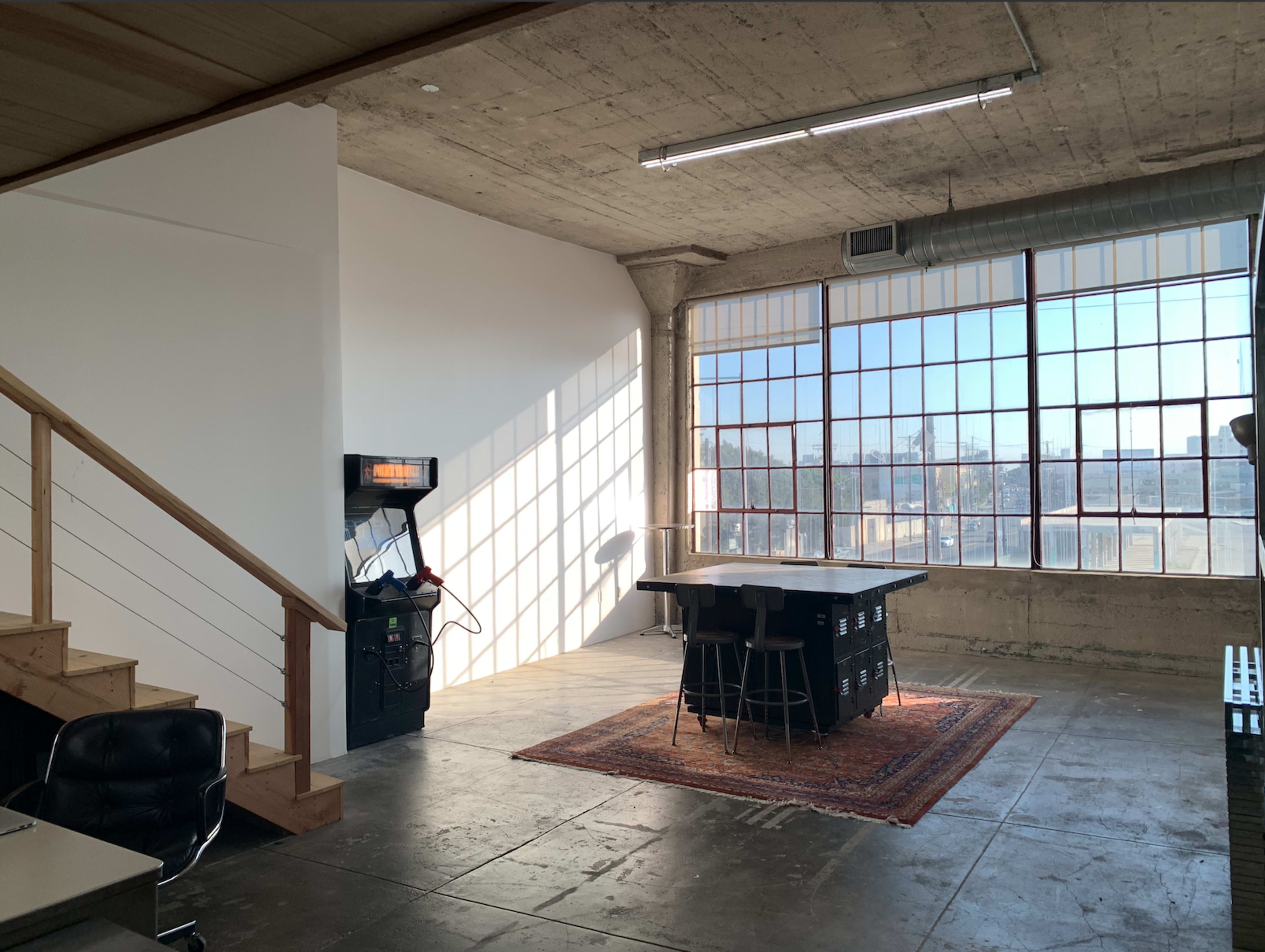 A spacious, industrial-style room with large windows, a staircase, a game machine, and a central table surrounded by stools on a patterned rug.