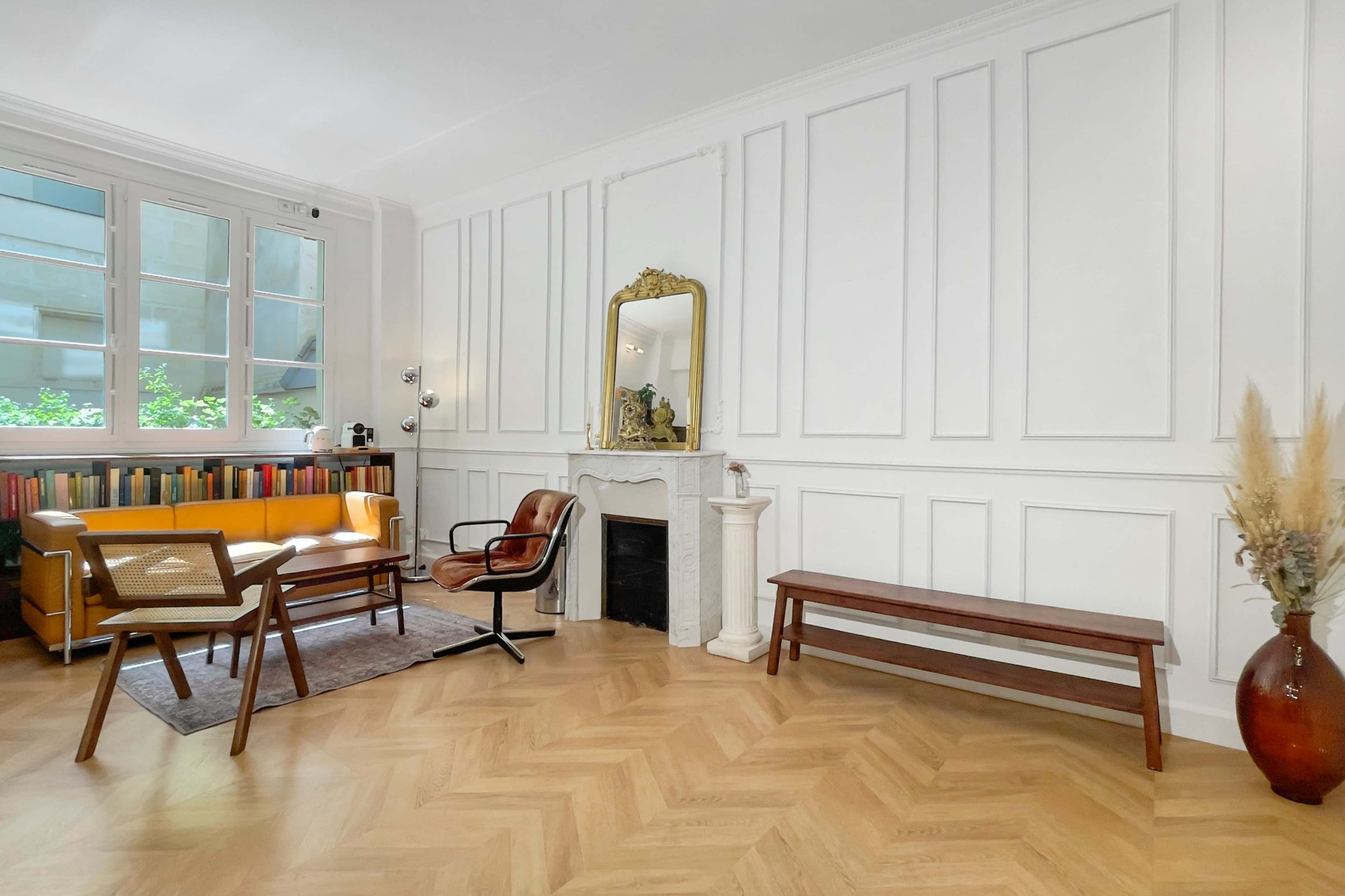 The image shows a living room with a modern chair and sofa, a wooden bench, a fireplace, and a large mirror against a wall with white paneling.