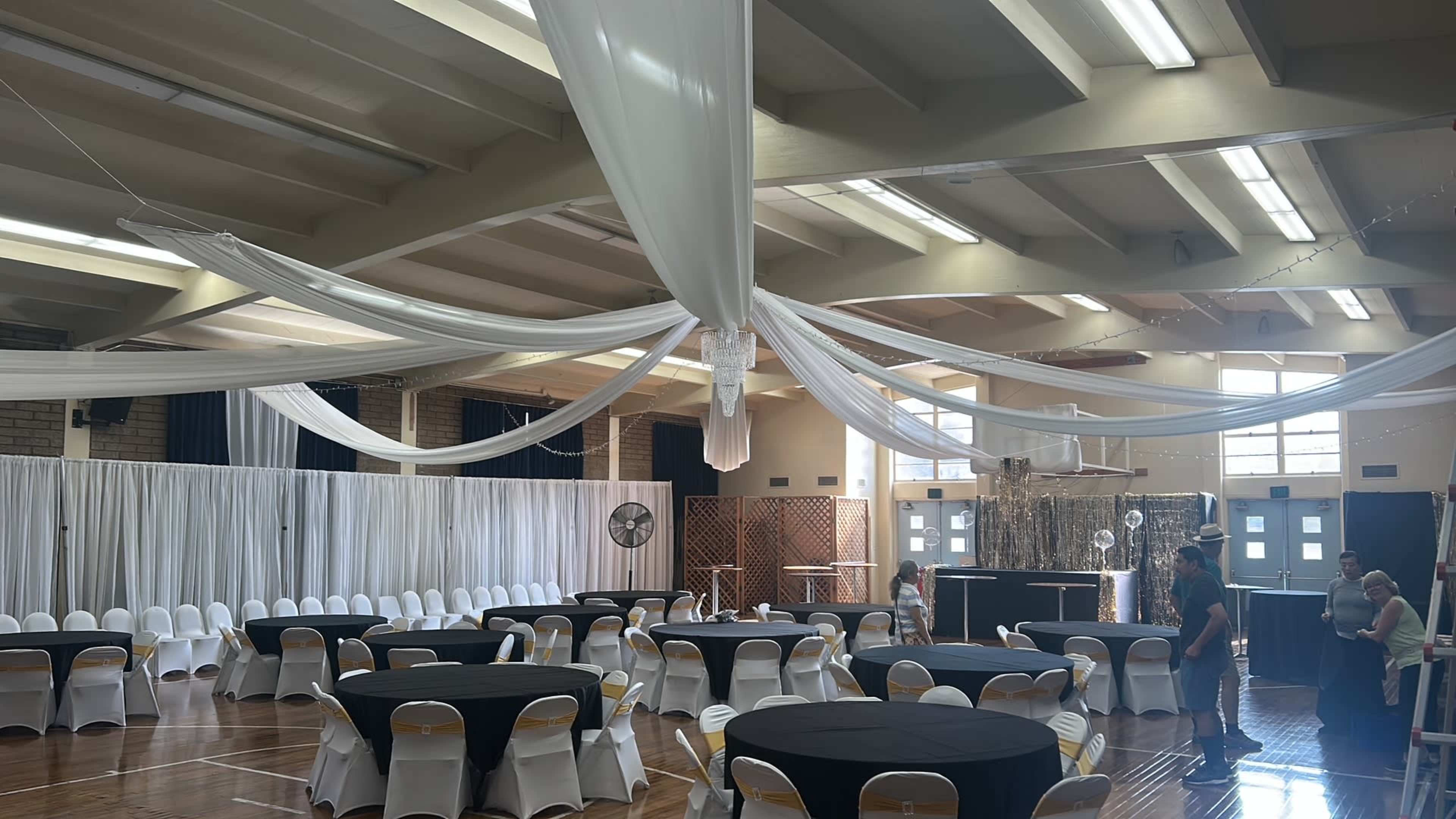 The image shows a spacious event hall decorated with draped white fabric overhead, black round tables with white chairs set up, and a shimmering backdrop at one end of the room.
