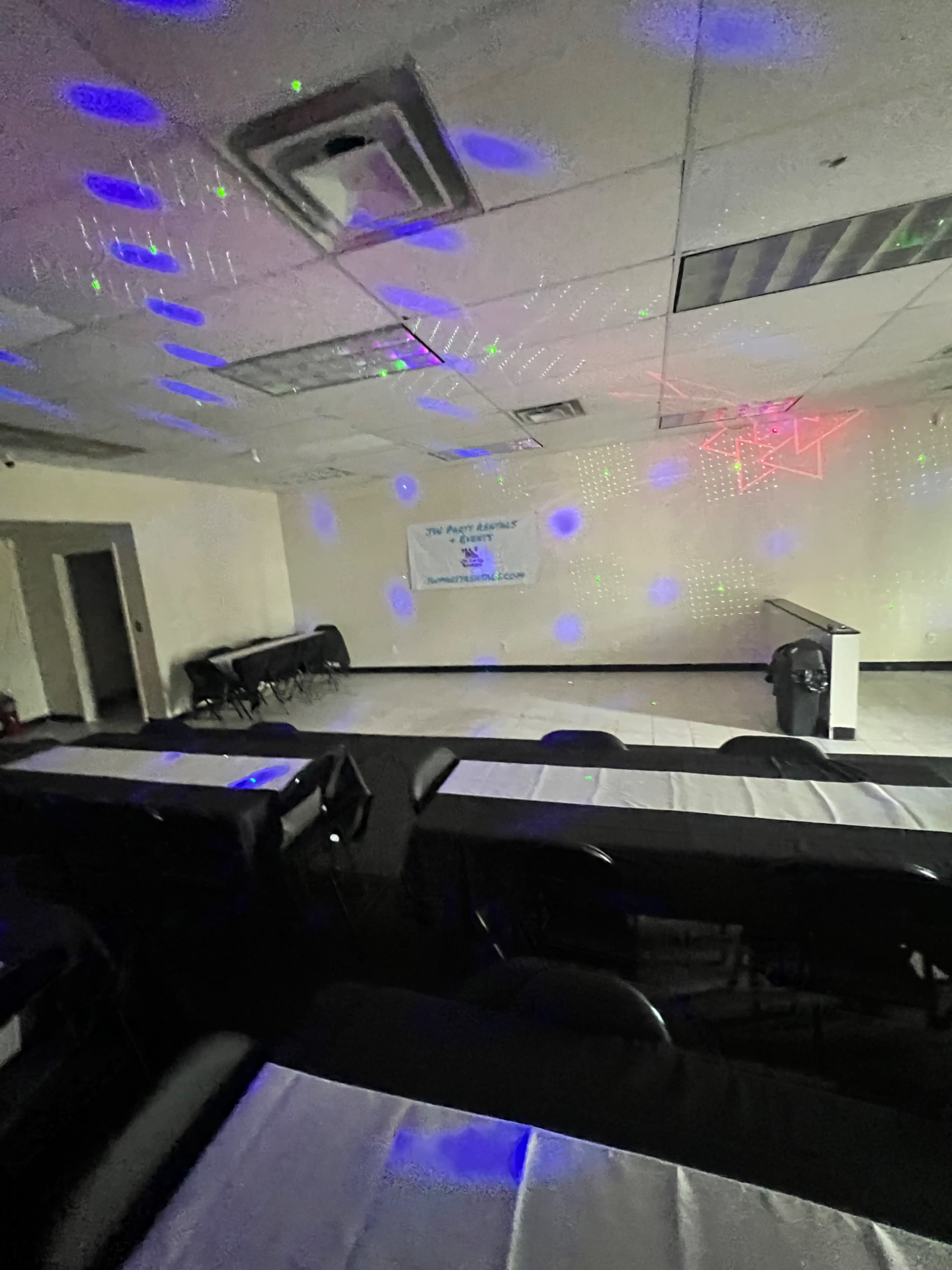The image shows a function room with rows of tables covered in white tablecloths, lit by colorful lights projecting patterns on the walls and ceiling.