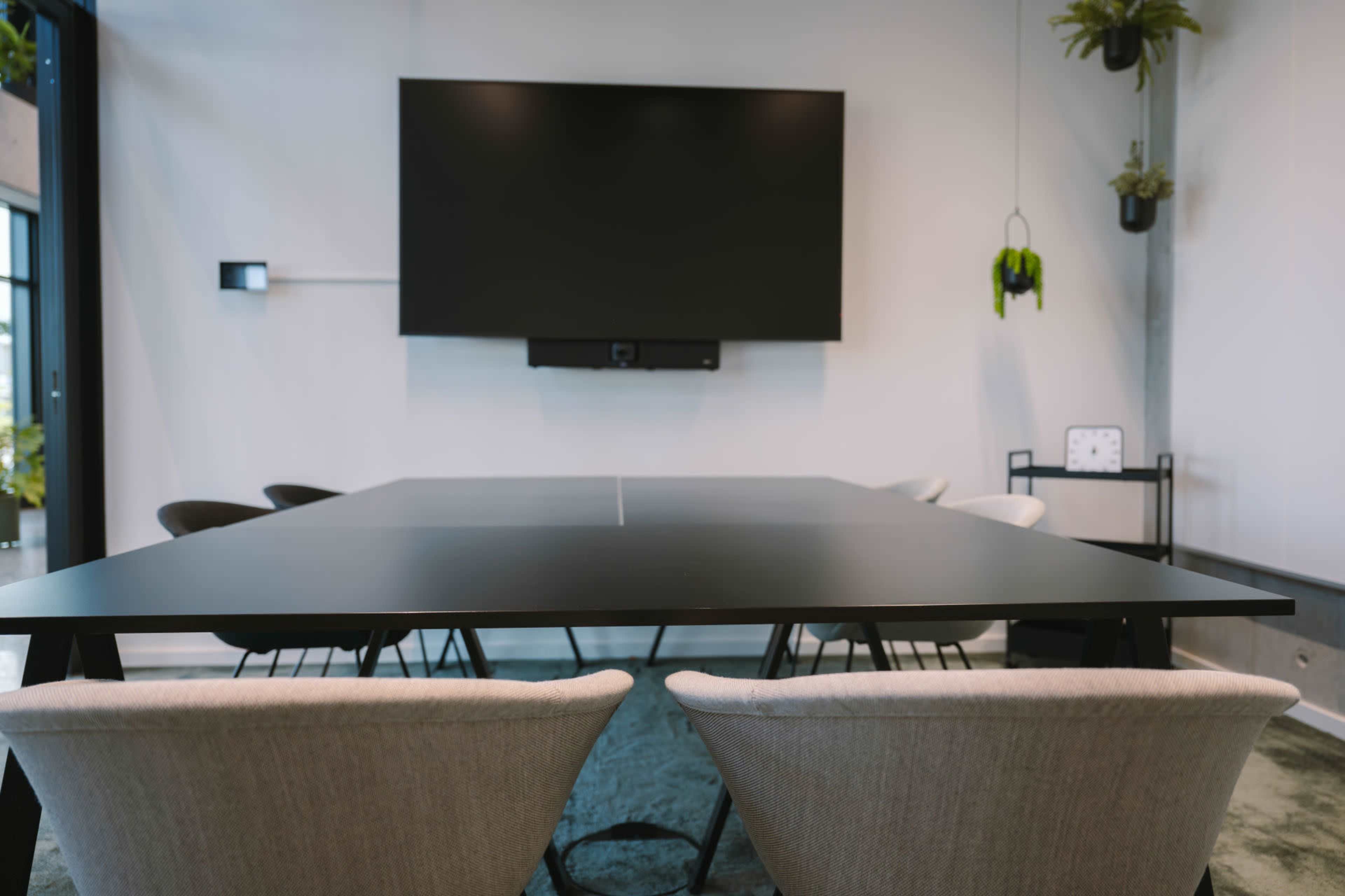 A modern conference room features a large black table, several chairs, a wall-mounted screen, and decorative plants.