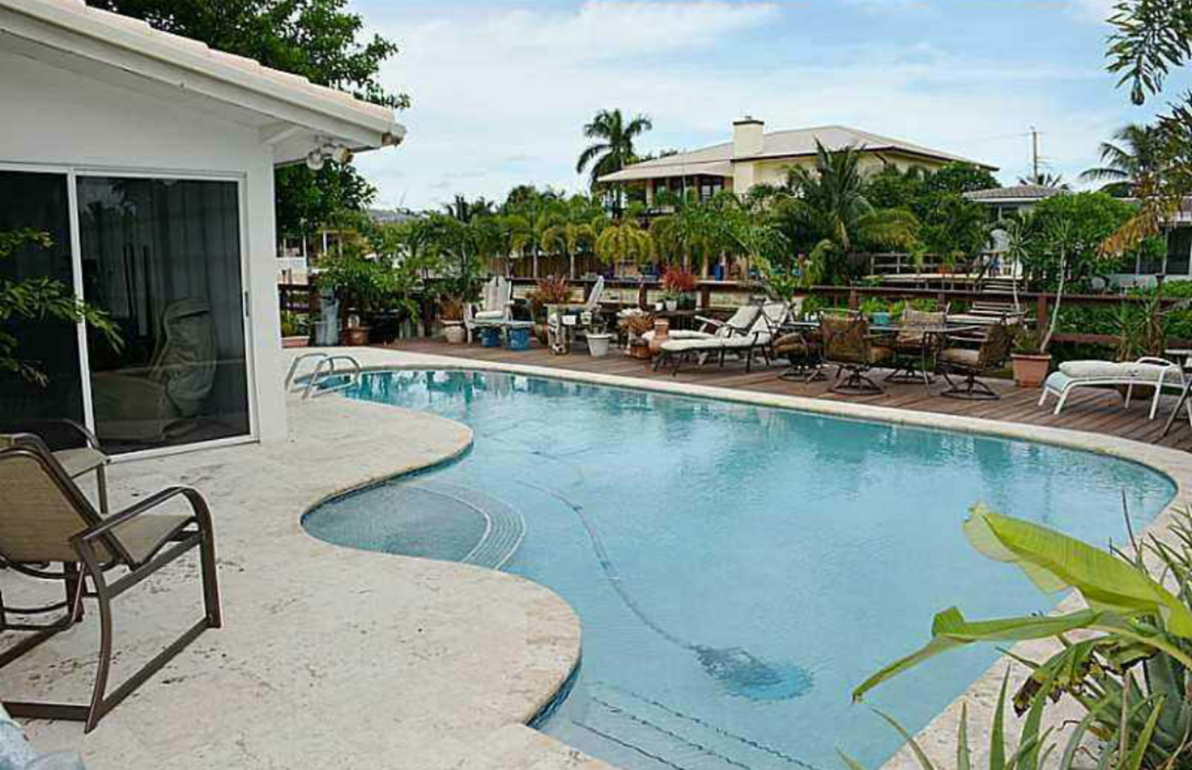 A swimming pool with a curved edge is surrounded by lounge chairs and tropical plants, adjacent to a house and palm trees.