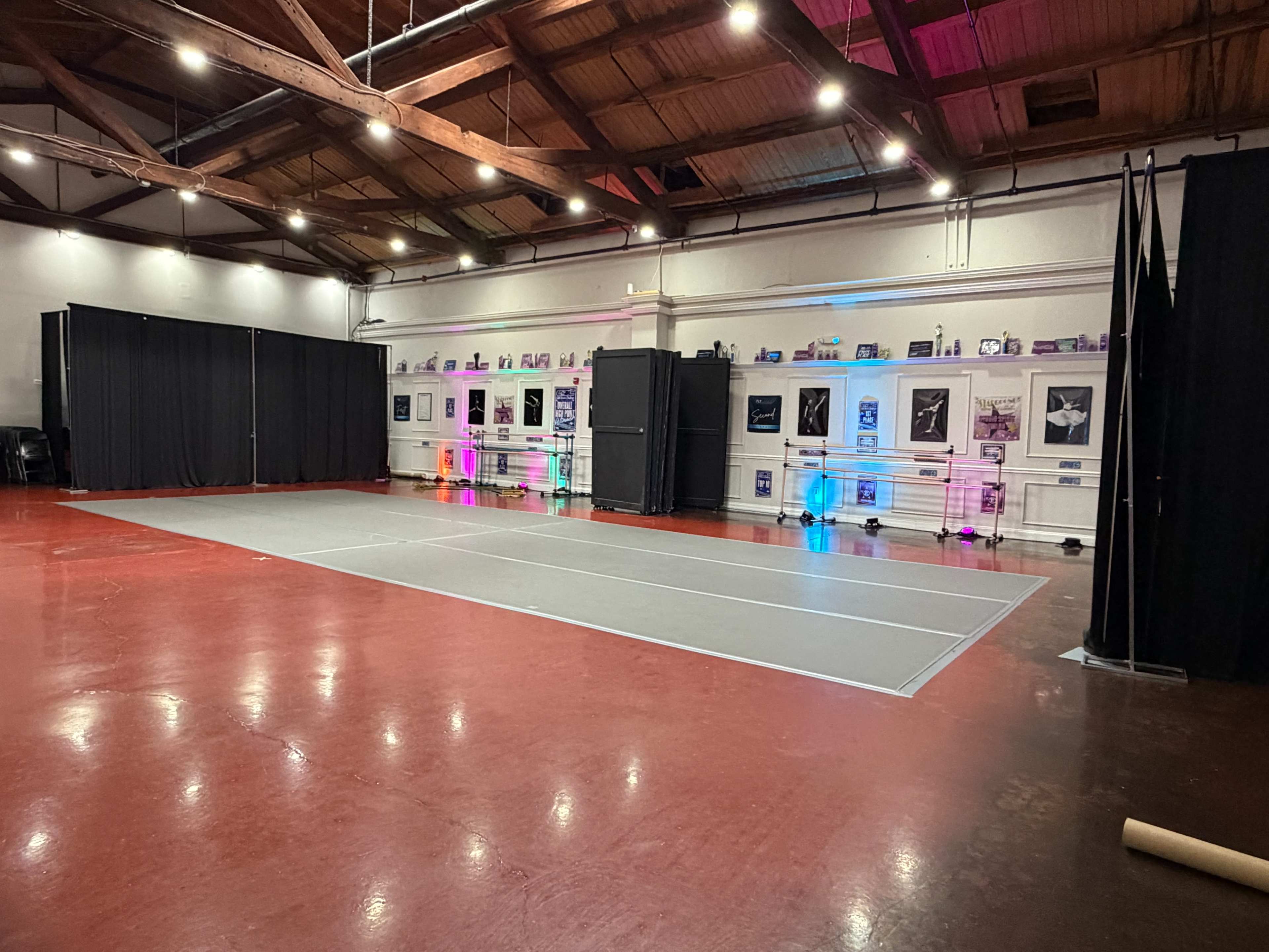 The image shows a spacious interior room with a gray dance floor, surrounded by black curtains and illuminated by colored lights, displaying various artworks on the walls.