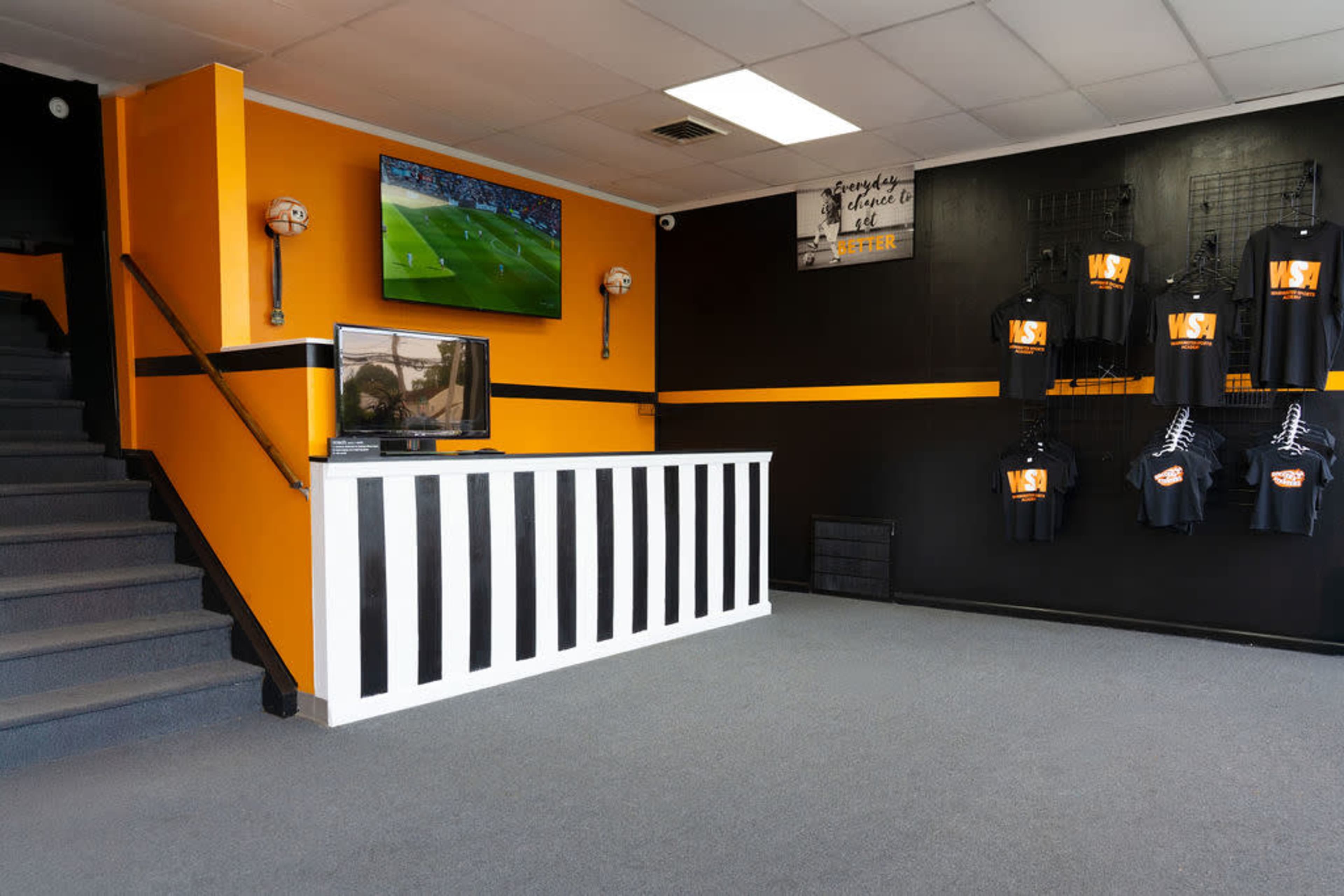 The interior of a shop features a reception area with a white and black striped counter, a TV displaying a sports game, and orange and black walls adorned with T-shirts hanging on display.