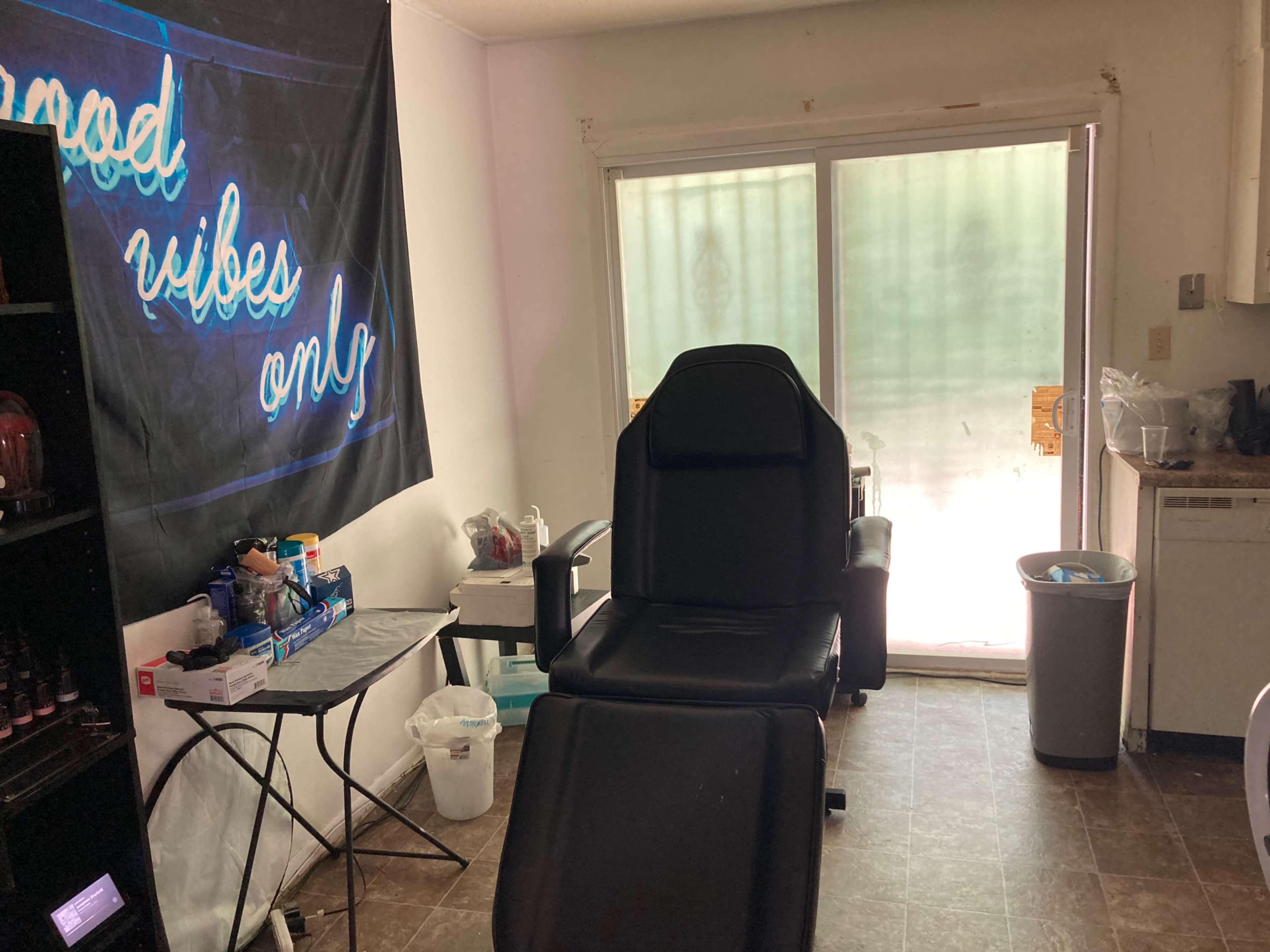 A room with a black tattoo chair, a small table with supplies, and a sliding glass door leading to an outdoor area.