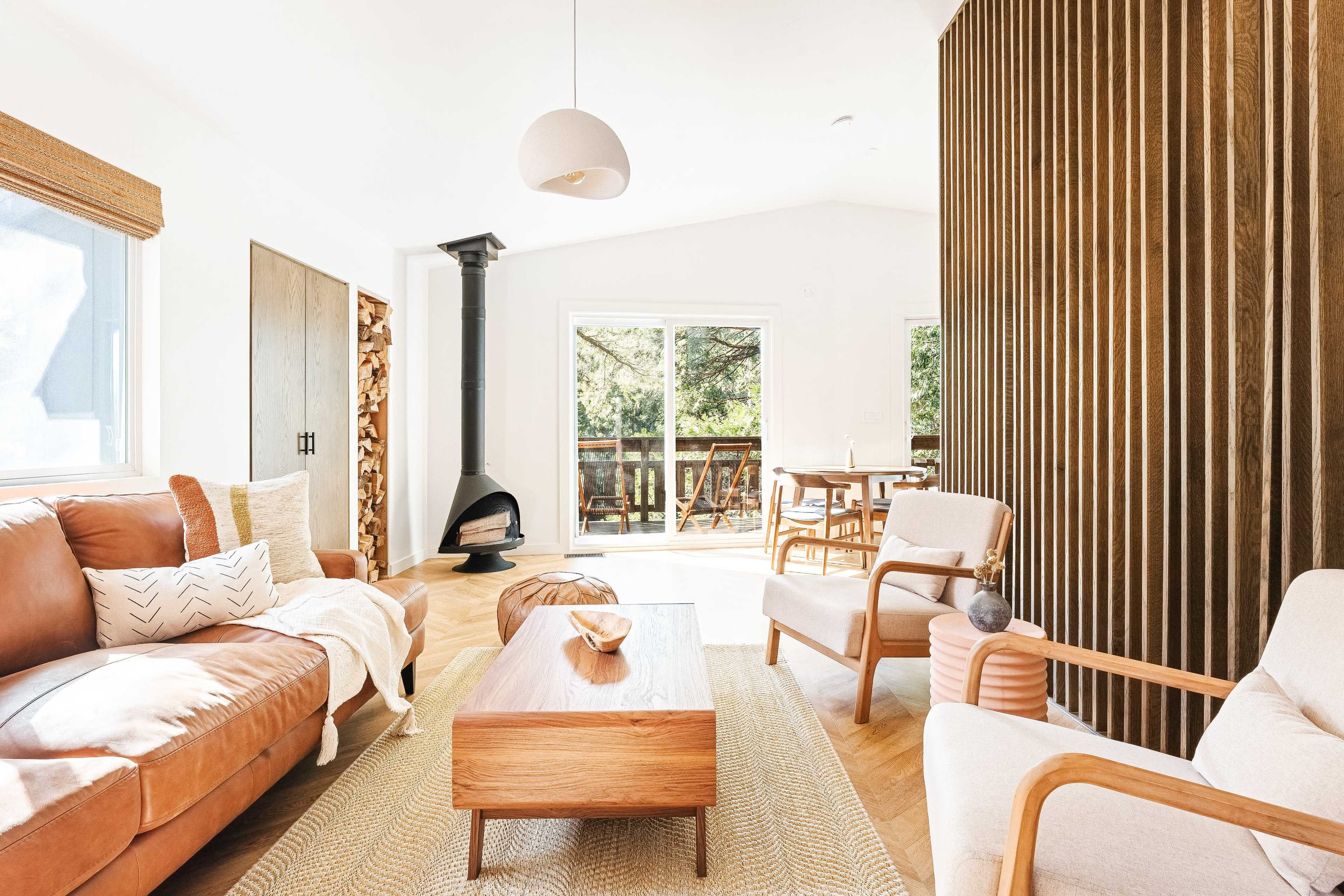 A modern living room features a brown leather sofa, a wooden coffee table, and a wood-burning stove, with large windows that overlook a deck surrounded by trees.