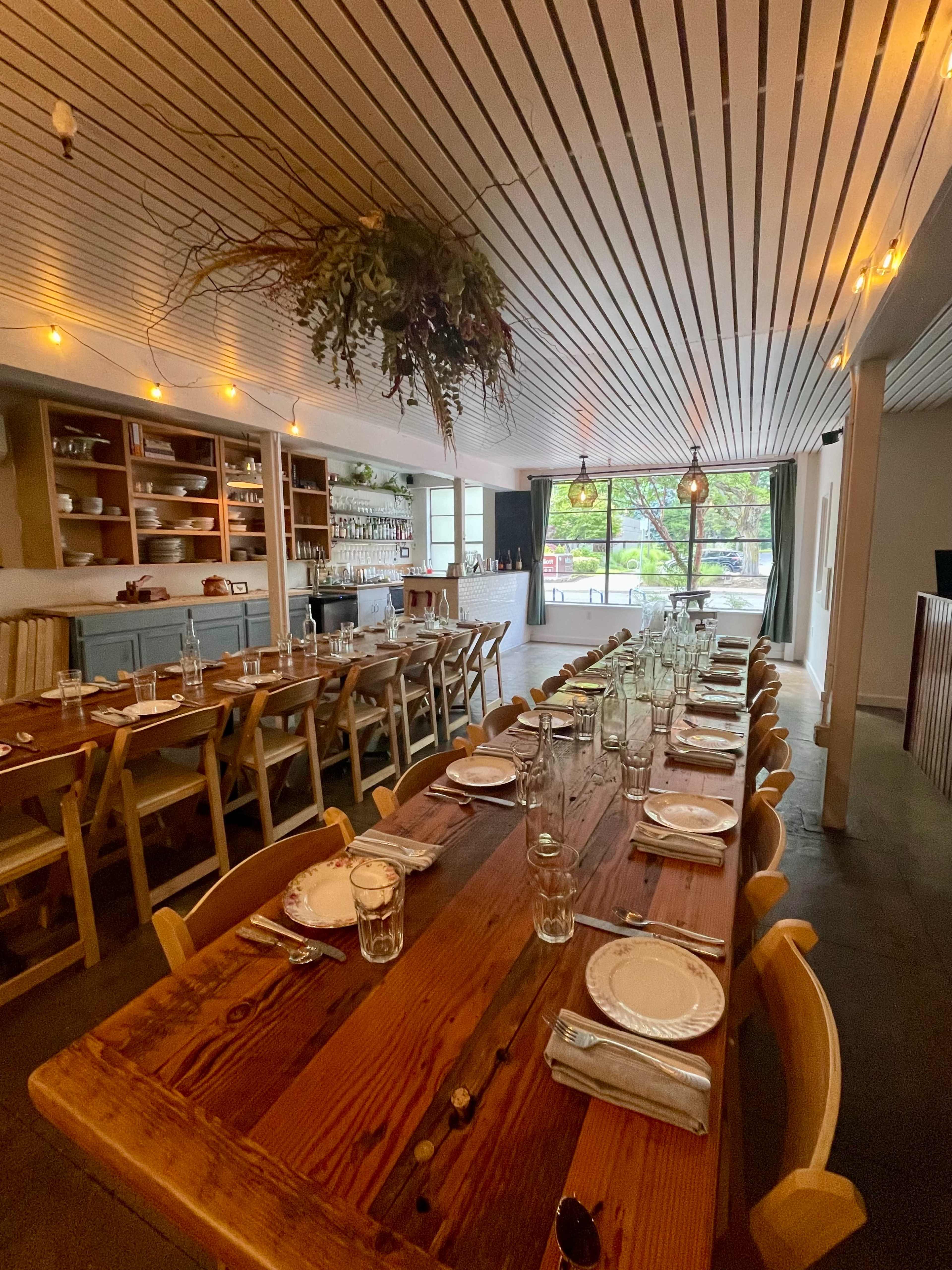 The image shows a spacious dining area with a long wooden table set for a meal, surrounded by chairs, and brightened by soft lighting and large windows.